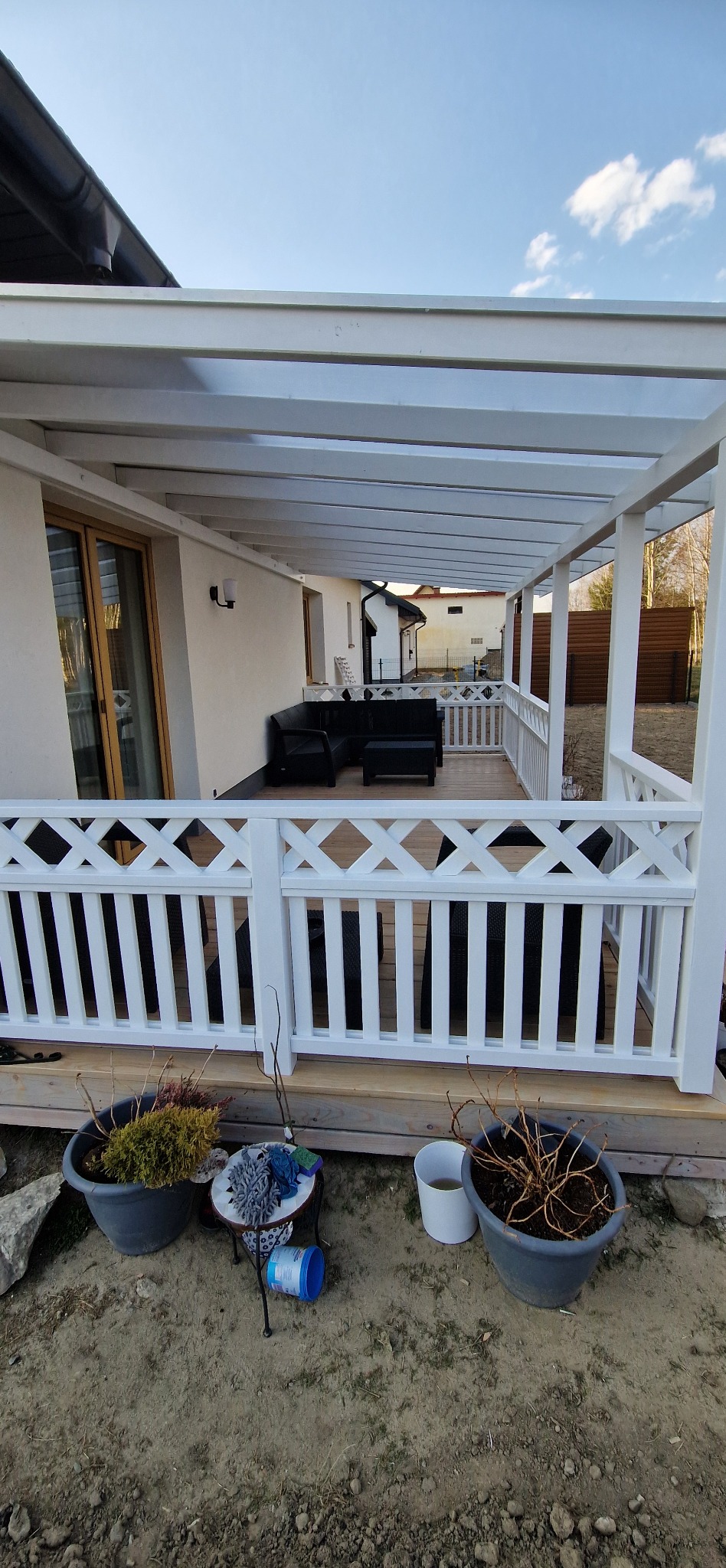 Biała altana z drewnianą podłogą i balustradą z motywem krzyża, na której stoi czarny komplet wypoczynkowy. Widok na ogród i dom.