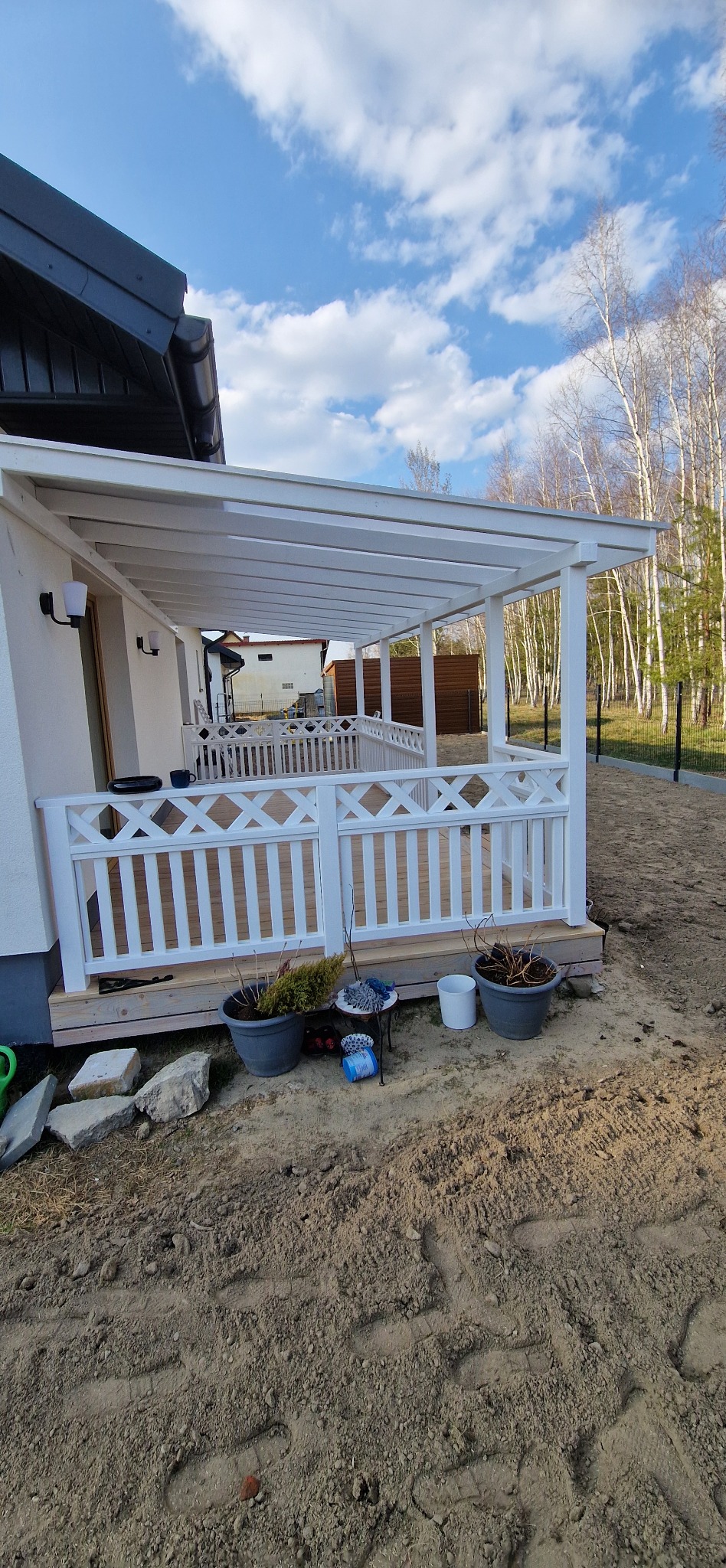 Biały taras z zadaszeniem przy domu, balustrada z motywem rombów. Widoczne donice i elementy dekoracyjne. W tle drzewa i błękitne niebo z chmurami.