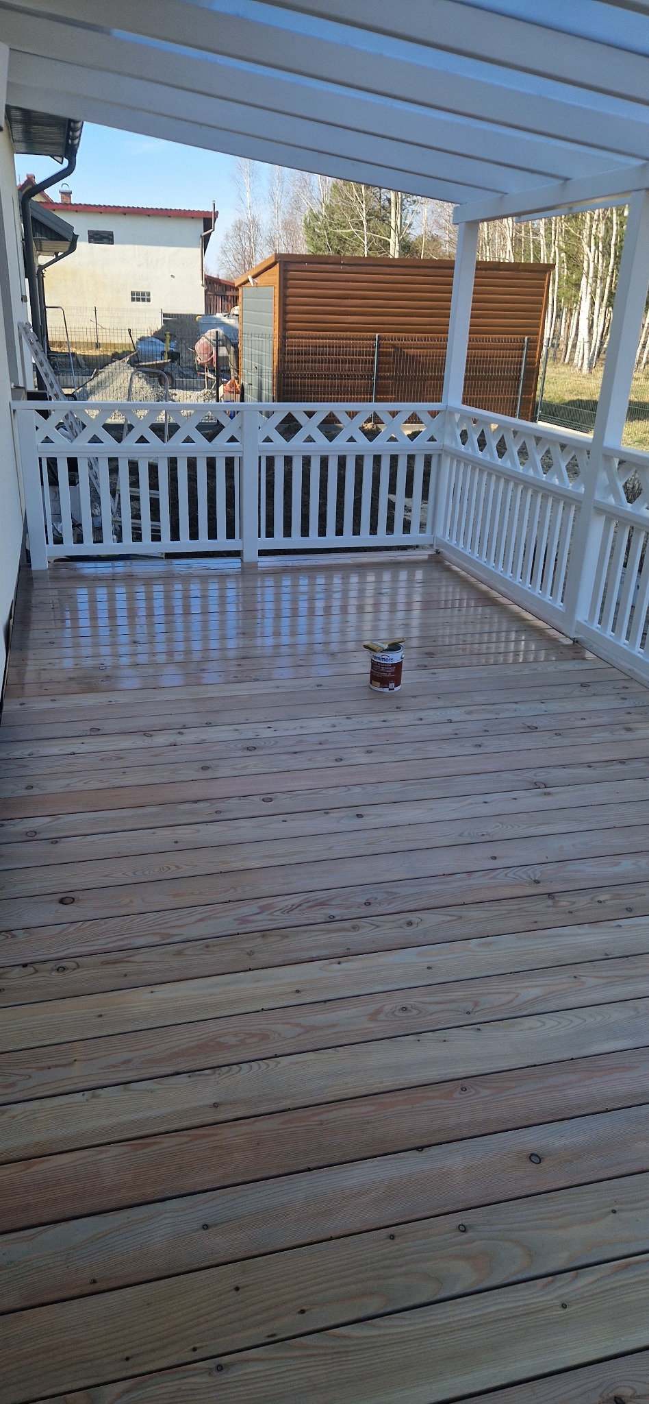 Świeżo polakierowany drewniany taras z białą balustradą. Na deskach stoi puszka z lakierem i pędzel. Widok na dom i budynek gospodarczy w tle, w słoneczny dzień.