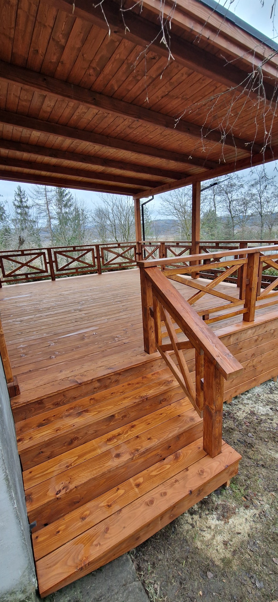 Zadaszony taras drewniany z balustradą krzyżową, widok na zewnątrz. Ciepłe oświetlenie podkreśla strukturę drewna. Widok na las w tle.