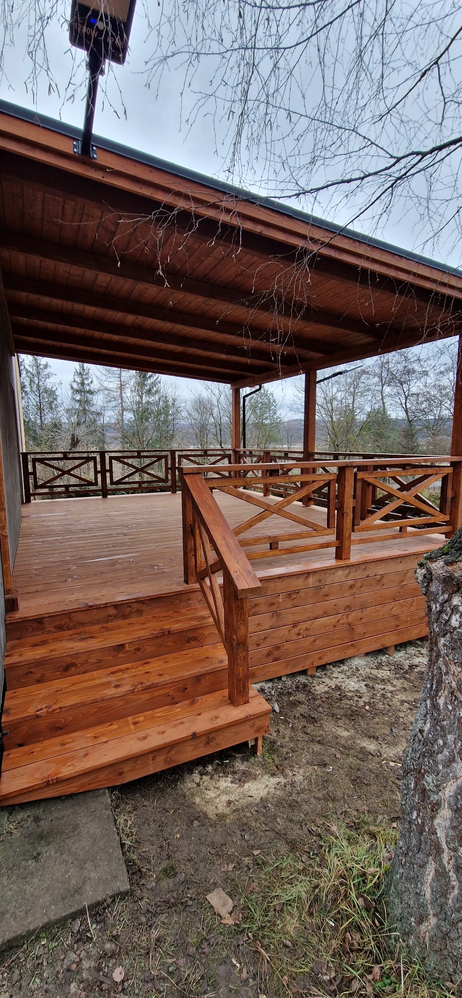 Zadaszony taras z drewna z balustradą krzyżową i schodami. Widoczne drzewa w tle. Oświetlenie solarne na dachu. Całość w ciepłym odcieniu brązu.