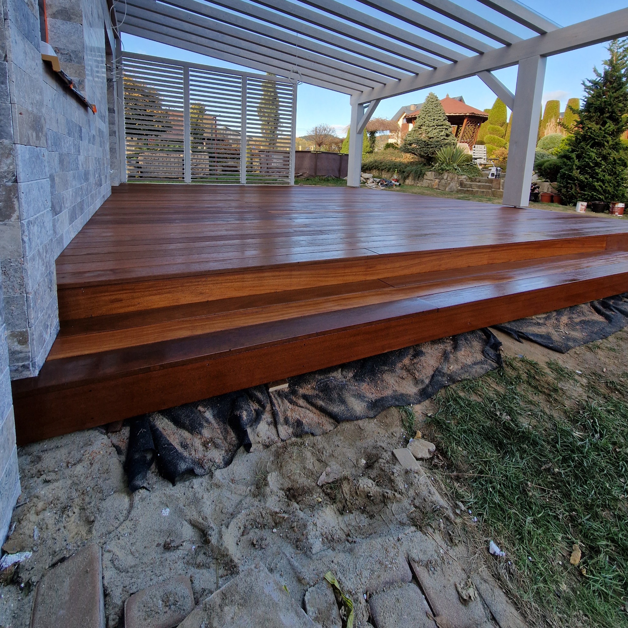 Nowy taras z drewna egzotycznego z niskimi schodami, połyskującą powierzchnią i pergolą. W tle widoczny ogród z altaną.