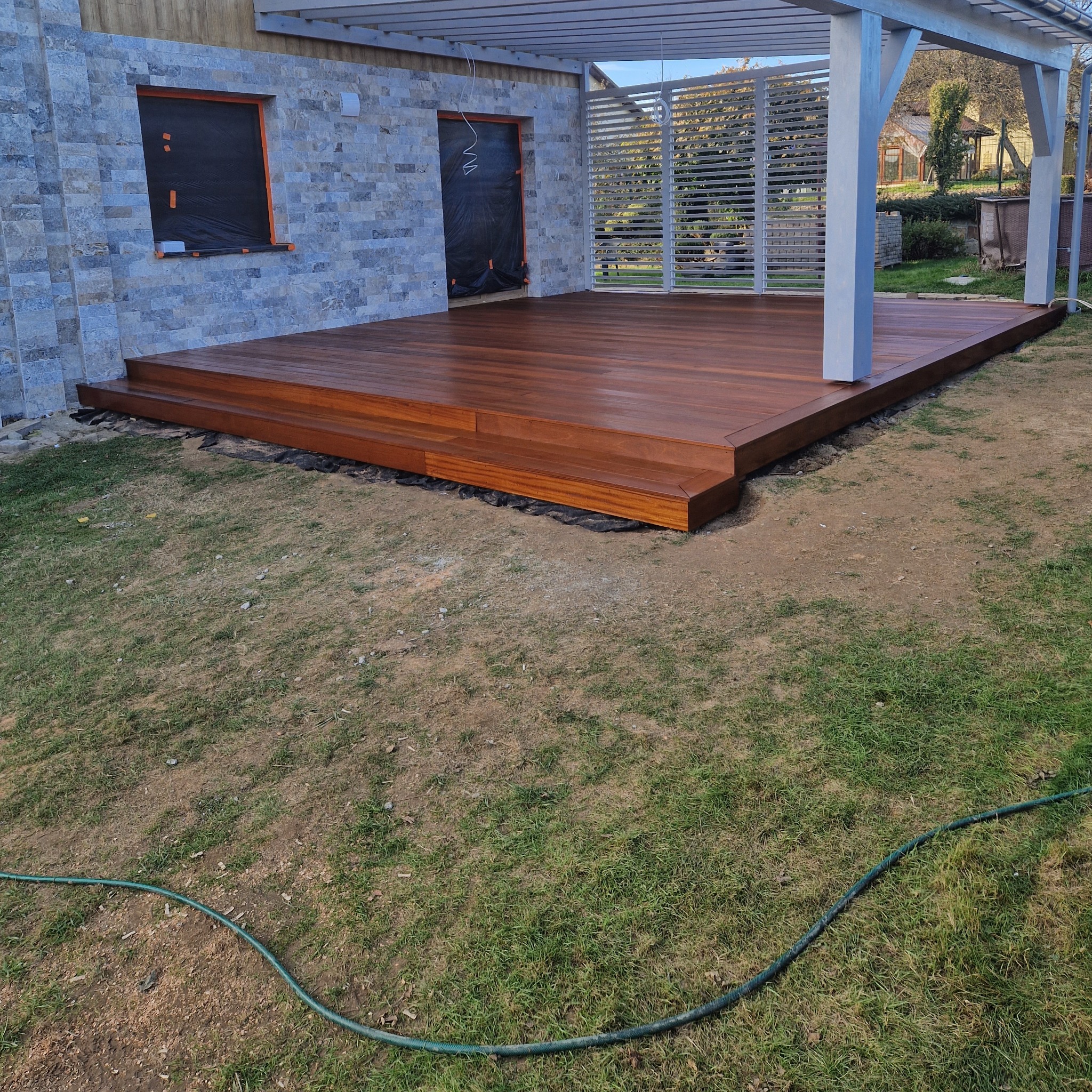 Elegancki taras drewniany z dwustopniowymi schodami, przylegający do budynku z kamienną elewacją i pergolą. Widoczny fragment trawnika i węża ogrodowego.