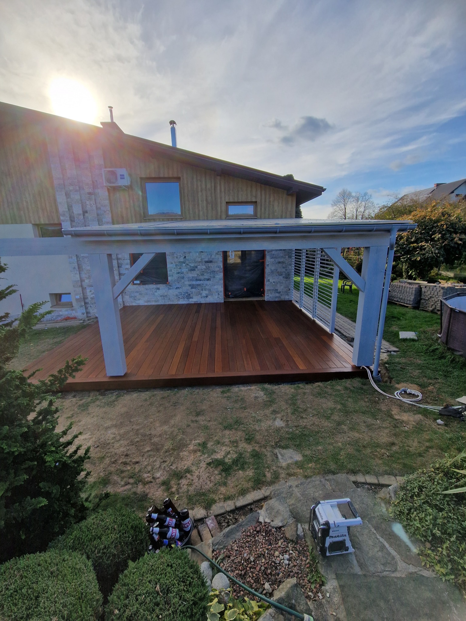 Nowoczesny taras z drewna egzotycznego, zadaszony pergolą w kolorze szarym, przylegający do domu z elementami kamienia. Widoczne narzędzia i butelki w tle.