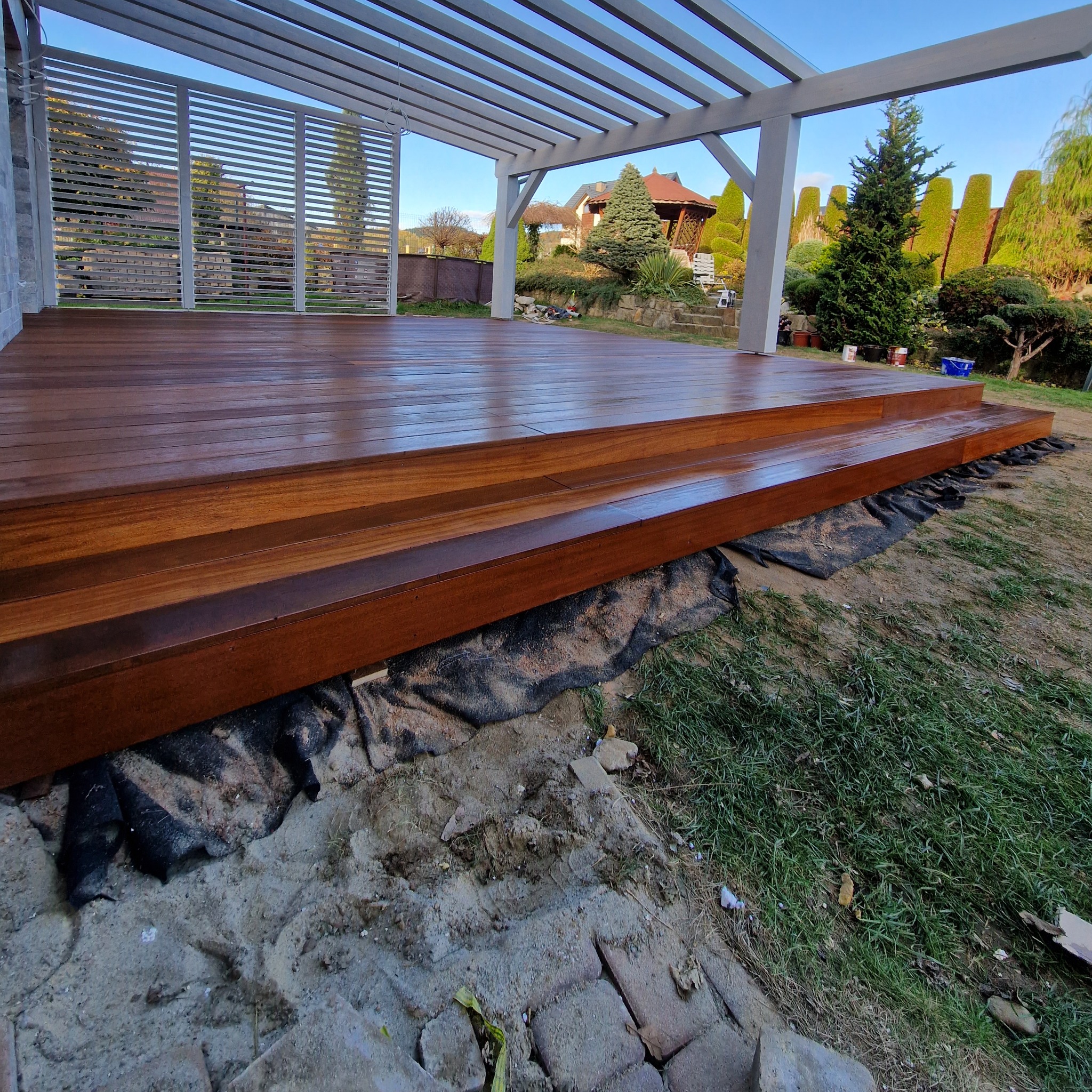 Drewniany taras z pergolą w ogrodzie. Schodki z drewna prowadzą na podest. Widoczne elementy konstrukcyjne i wykończenie drewna. W tle zieleń i zabudowa ogrodowa.