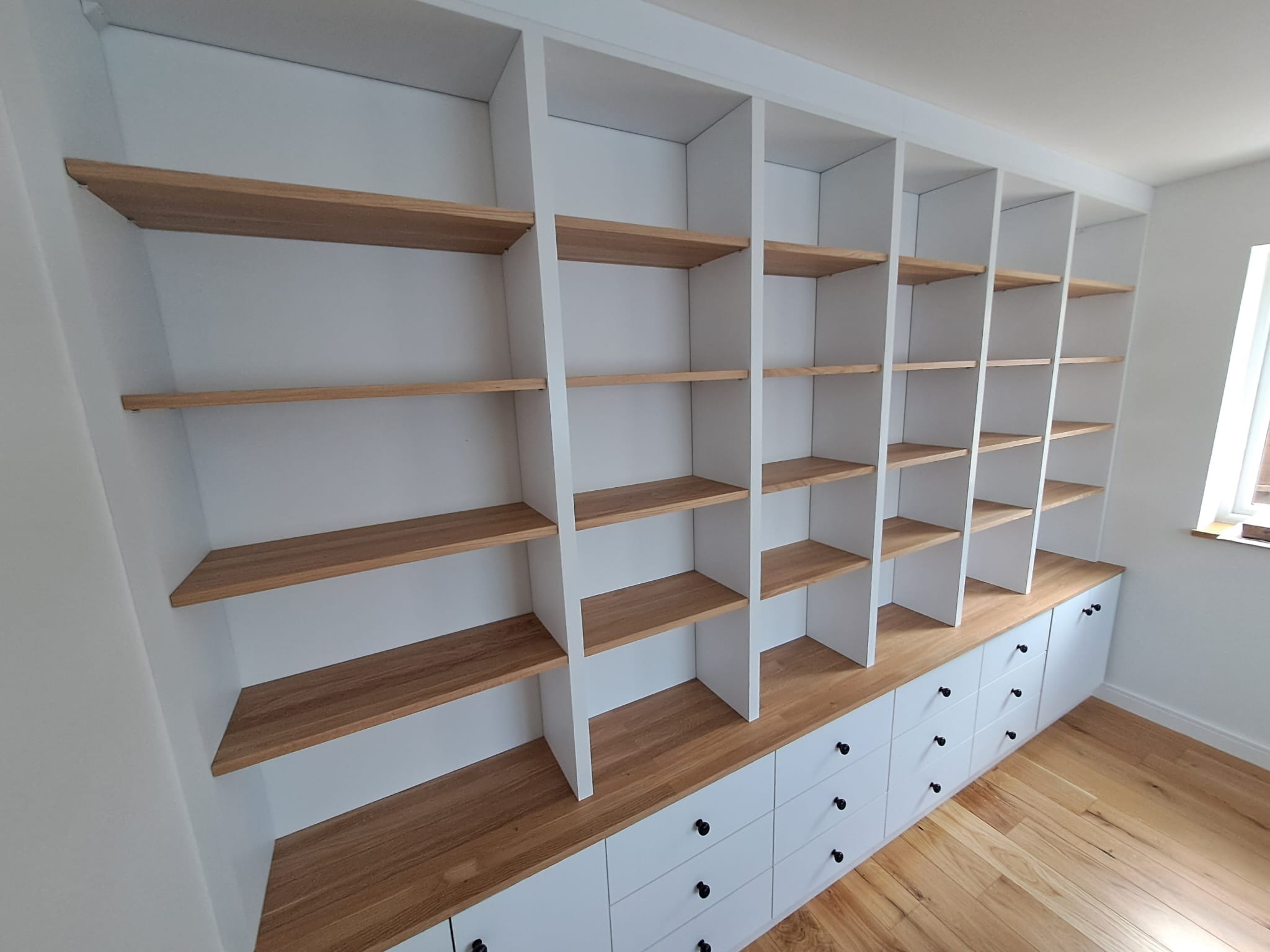 Biała zabudowa meblowa z drewnianymi półkami i szafkami z czarnymi uchwytami, wkomponowana w ścianę, z widoczną podłogą w odcieniach drewna.