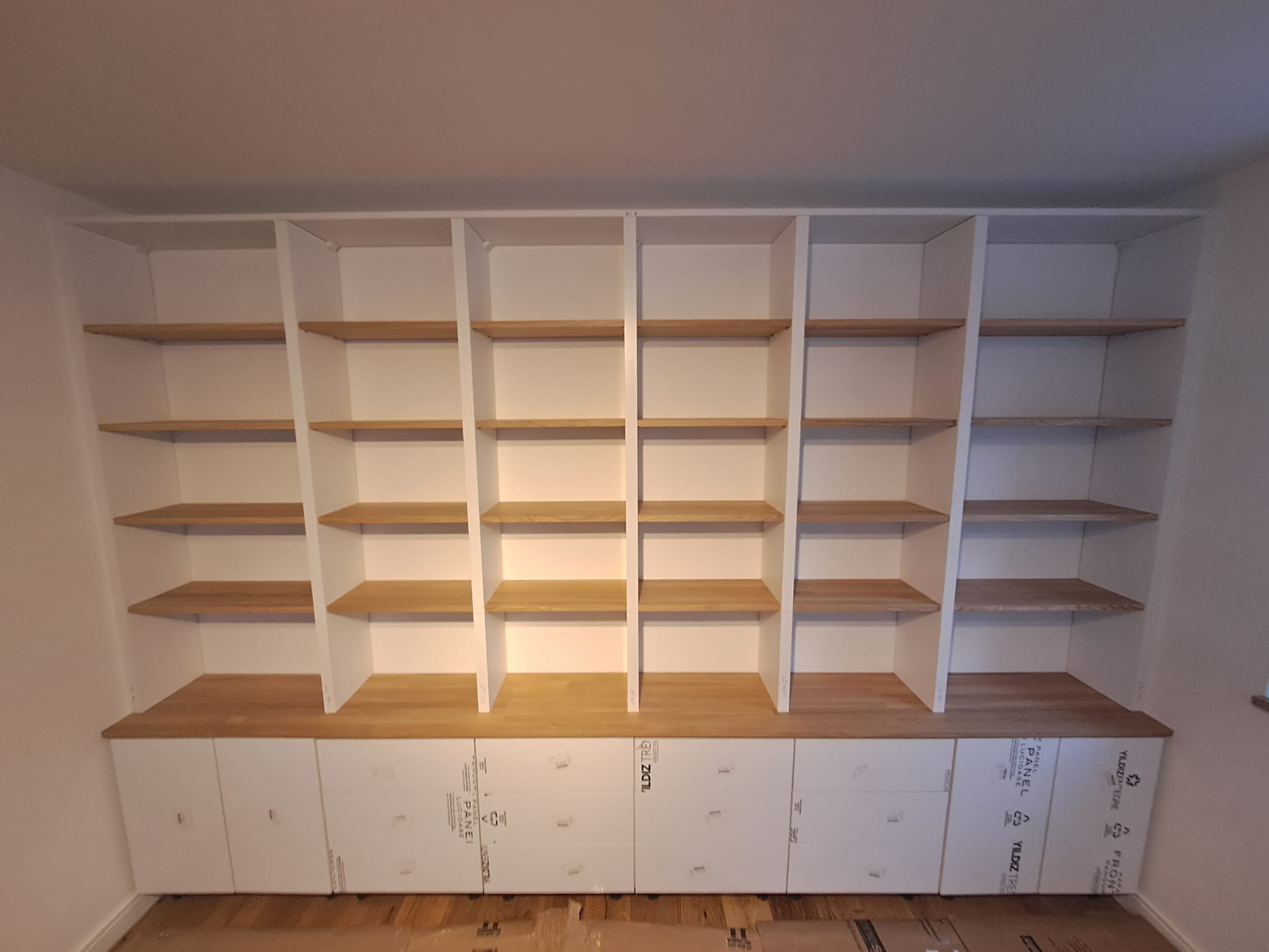 Biała zabudowa meblowa z drewnianymi półkami i szafkami z folią ochronną. Widok na regał wnękowy z otwartymi półkami i dolnymi szafkami.
