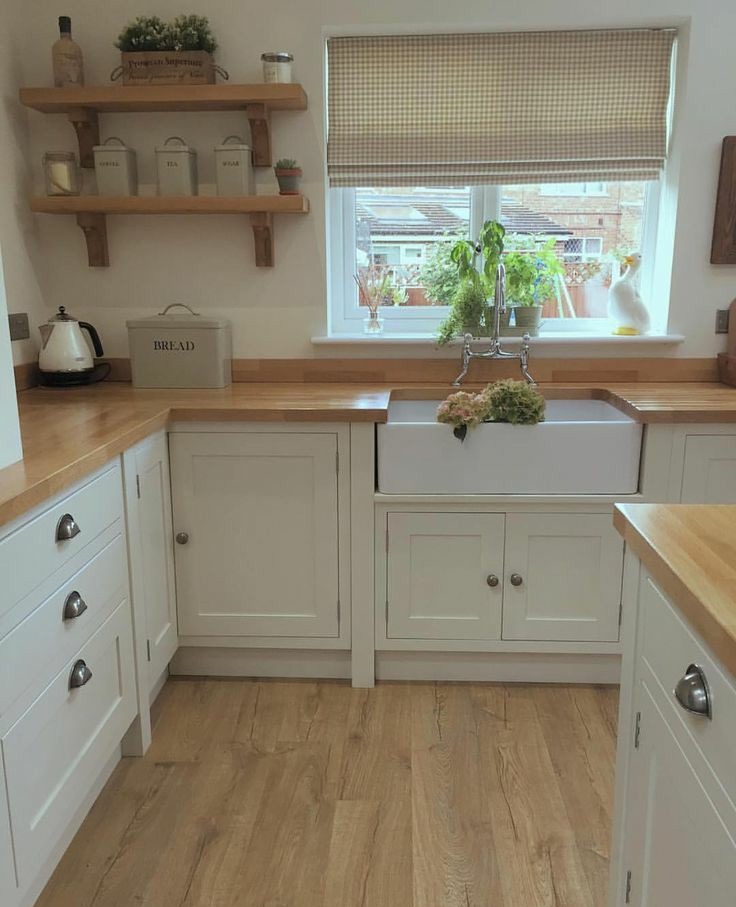 Jasna kuchnia w stylu rustykalnym: białe szafki z drewnianymi blatami, ceramiczny zlew pod oknem z roletą w kratę, drewniana podłoga i półki z dekoracjami.