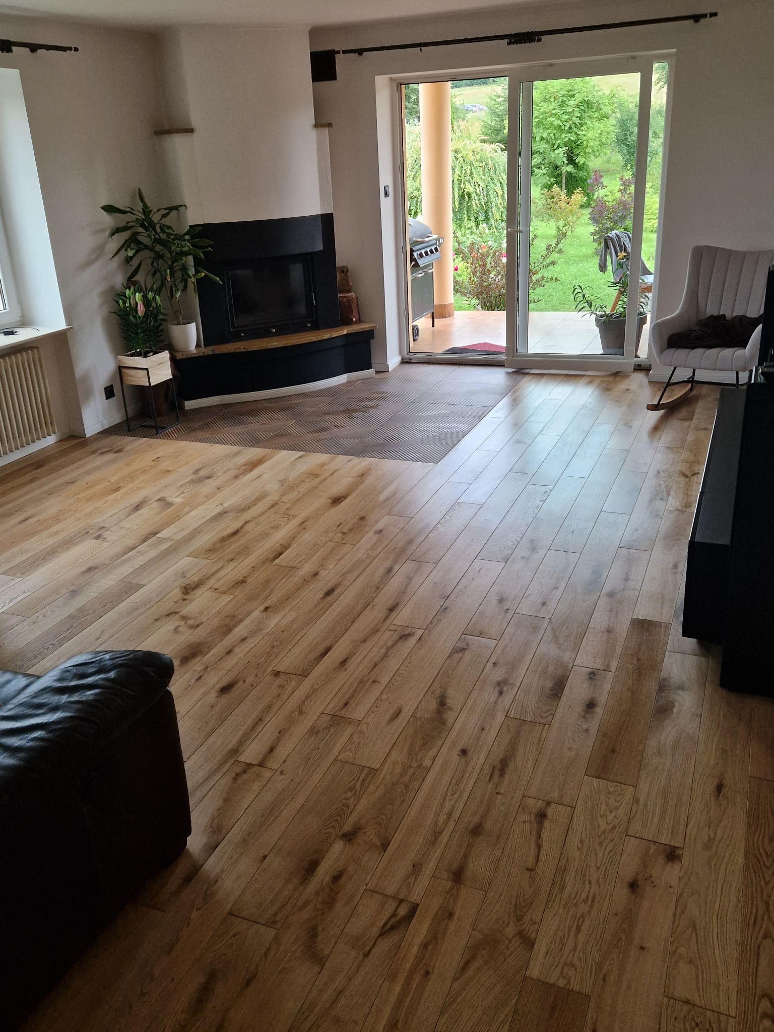 Jasne panele podłogowe z widoczną strukturą drewna w przestronnym salonie z kominkiem i wyjściem na taras, częściowo widoczny narożnik sofy.