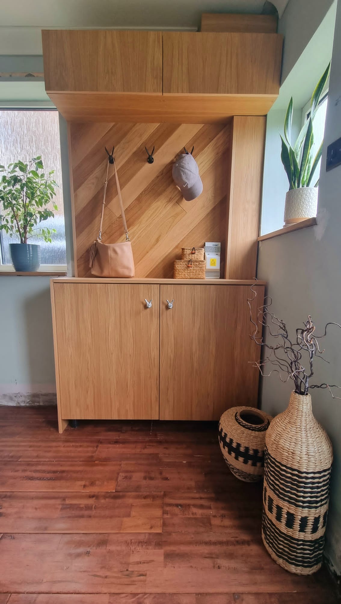 Drewniana szafa wnękowa z wieszakami i półkami, wykończona w jasnym drewnie, z dekoracyjnymi uchwytami. W tle widoczne okno i rośliny doniczkowe.