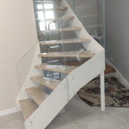 Piotr Tkacz Usługi Stolarskie - Nowoczesne schody z drewnianymi stopniami zabezpieczonymi folią i szklaną balustradą, biała konstrukcja nośna. Widok z dołu, jasne wnętrze.