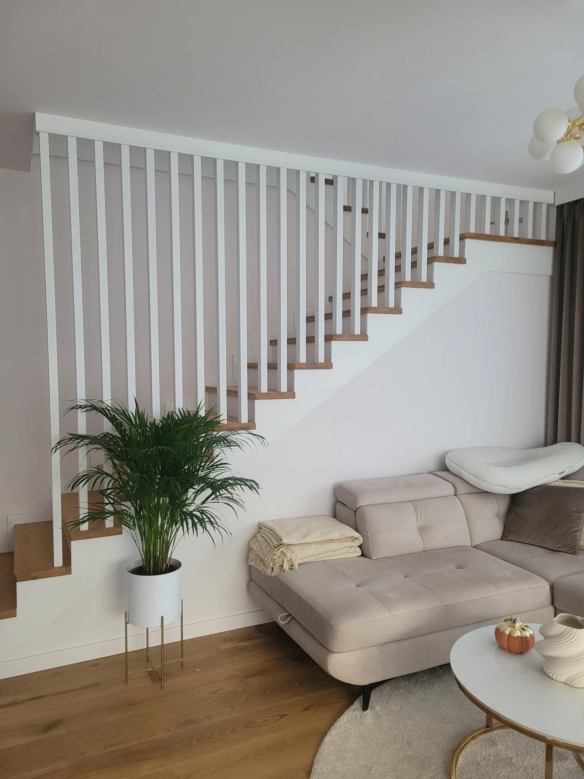Nowoczesne schody z drewnianymi stopniami i białą, minimalistyczną balustradą z pionowych listew, widoczne z perspektywy salonu z beżową sofą i rośliną doniczkową.