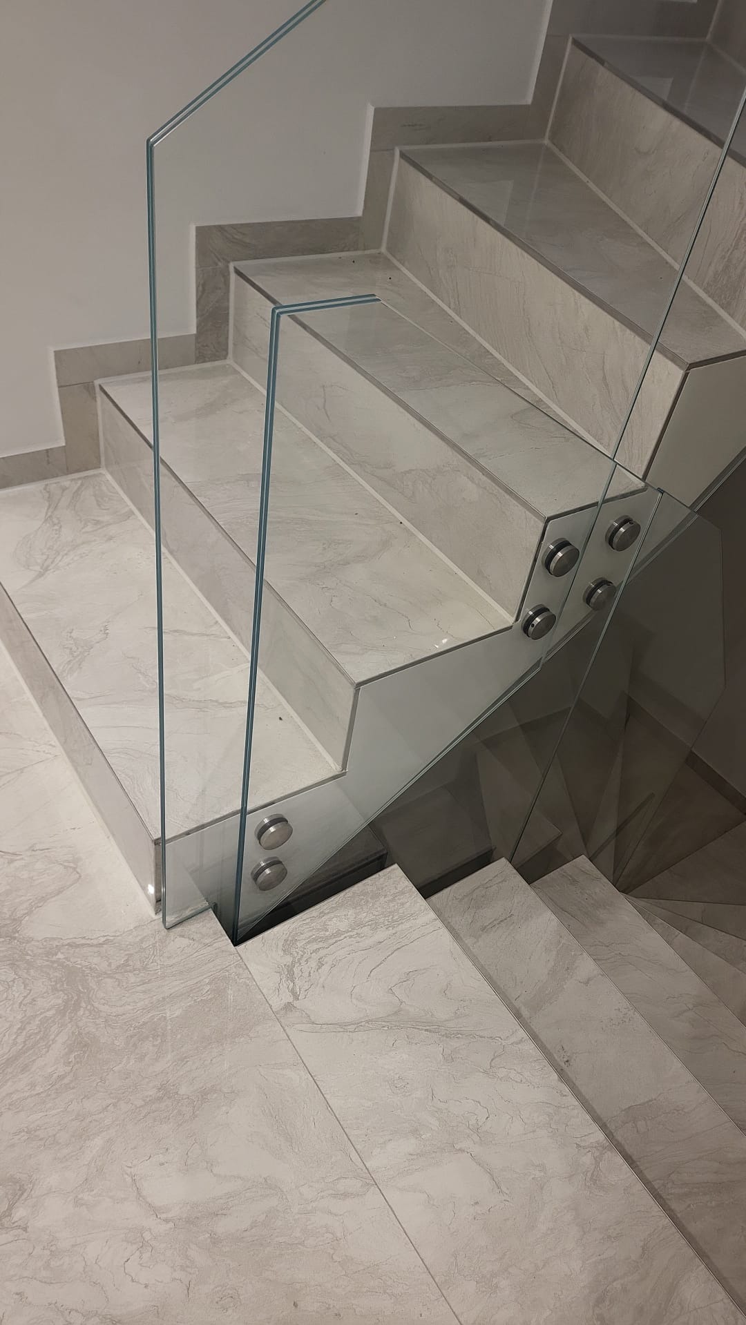 Nowoczesne schody z marmurowymi stopniami i szklaną balustradą mocowaną na metalowych uchwytach. Minimalistyczny design, jasna kolorystyka, widok z góry.