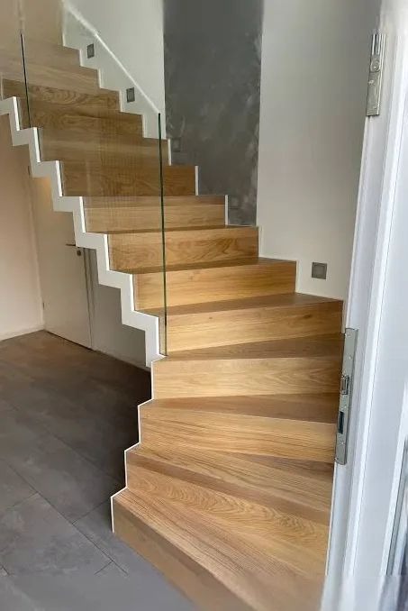 Nowoczesne schody drewniane z białymi podstopnicami i szklaną balustradą, widok z dołu, jasne drewno, minimalistyczny design, oświetlenie punktowe na ścianie.