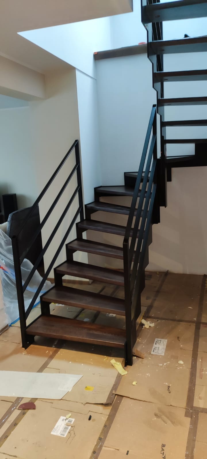 Nowoczesne schody z drewnianymi stopniami i czarną, minimalistyczną metalową balustradą. Widok z dołu, na podłodze karton zabezpieczający.