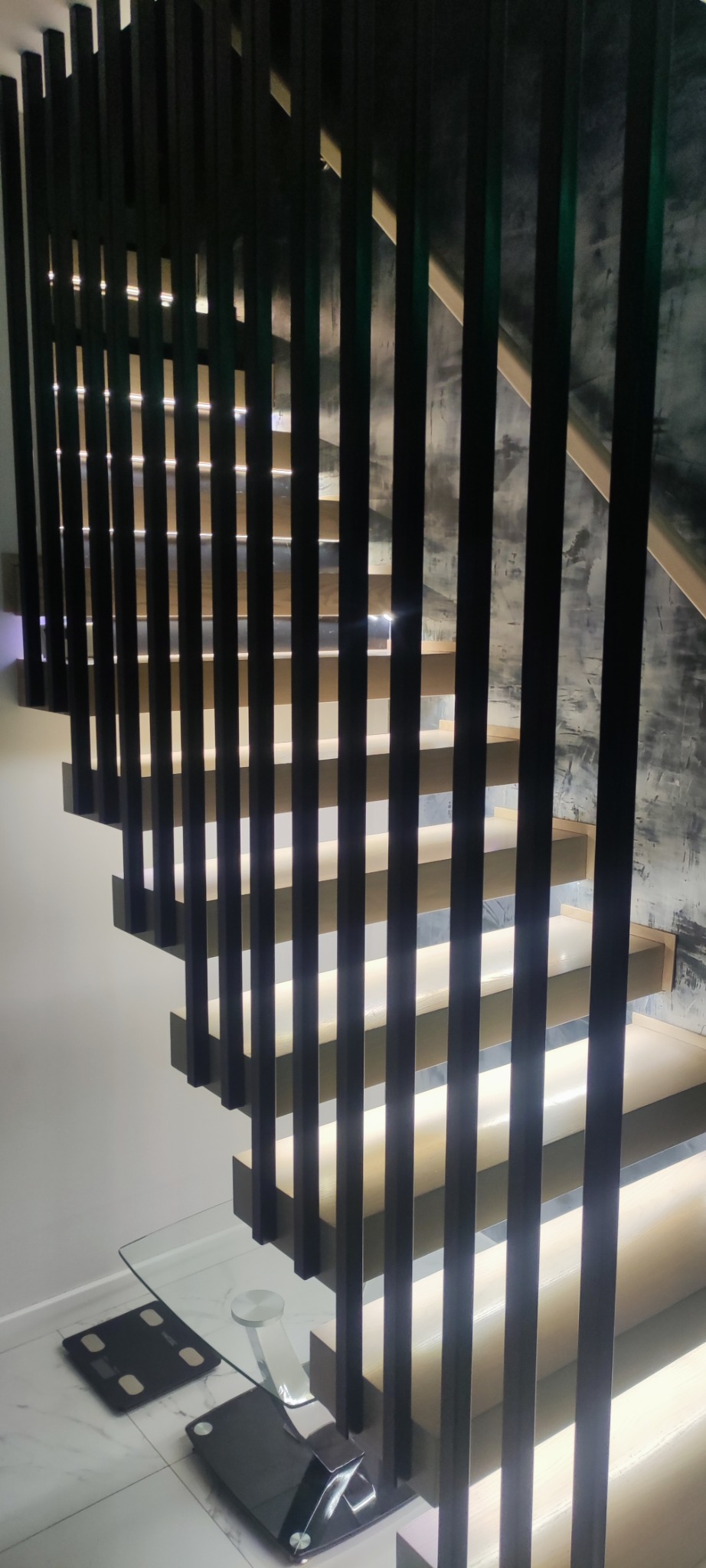 Nowoczesne schody z podświetlanymi stopniami i czarną balustradą z pionowych elementów. Widok z dołu, podkreślający minimalistyczny design i oświetlenie.