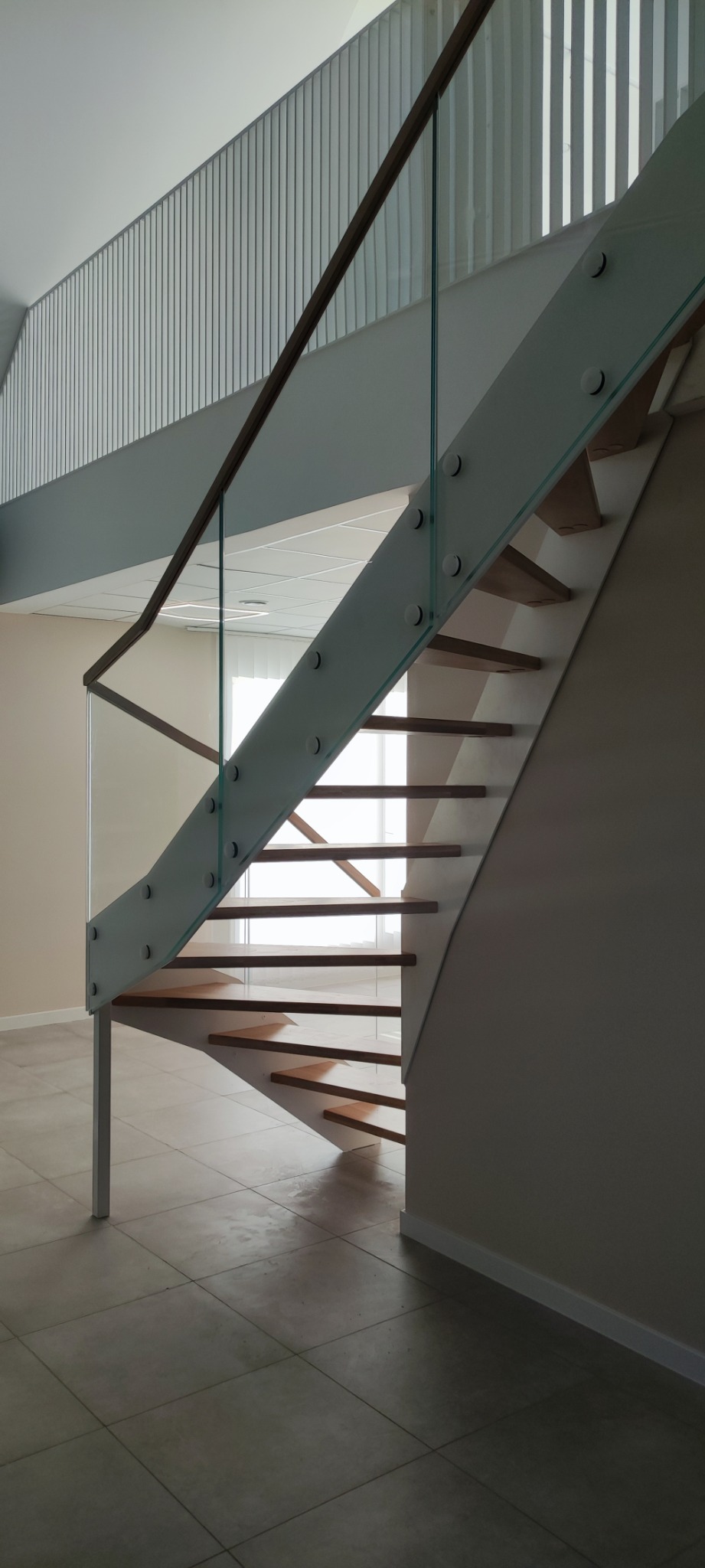 Nowoczesne schody z drewnianymi stopniami i szklaną balustradą, mocowaną na metalowych wspornikach. Widoczna barierka na antresoli z pionowych, białych elementów.