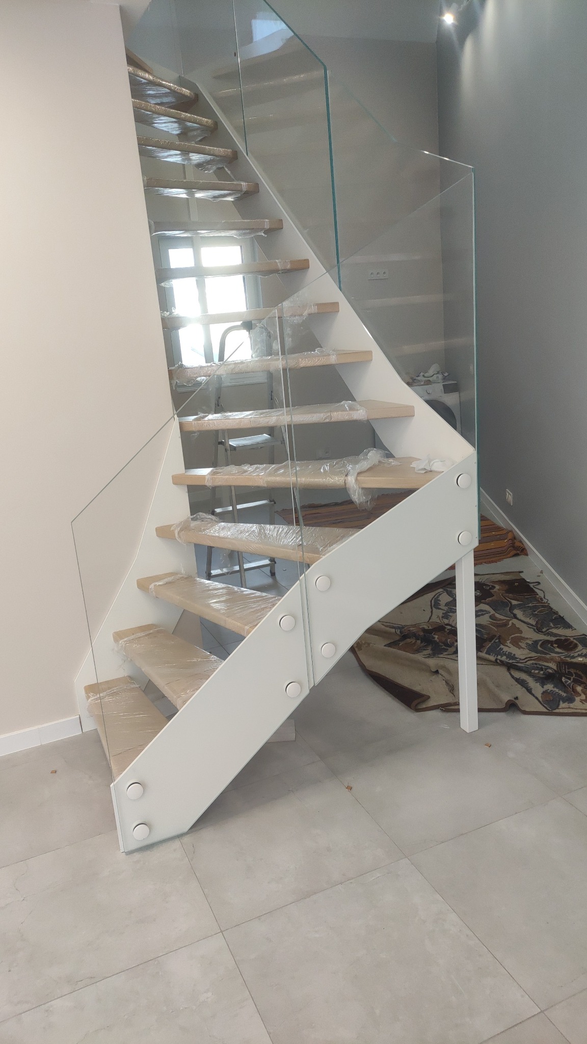 Nowoczesne schody z drewnianymi stopniami zabezpieczonymi folią i szklaną balustradą, biała konstrukcja nośna. Widok z dołu, jasne wnętrze.
