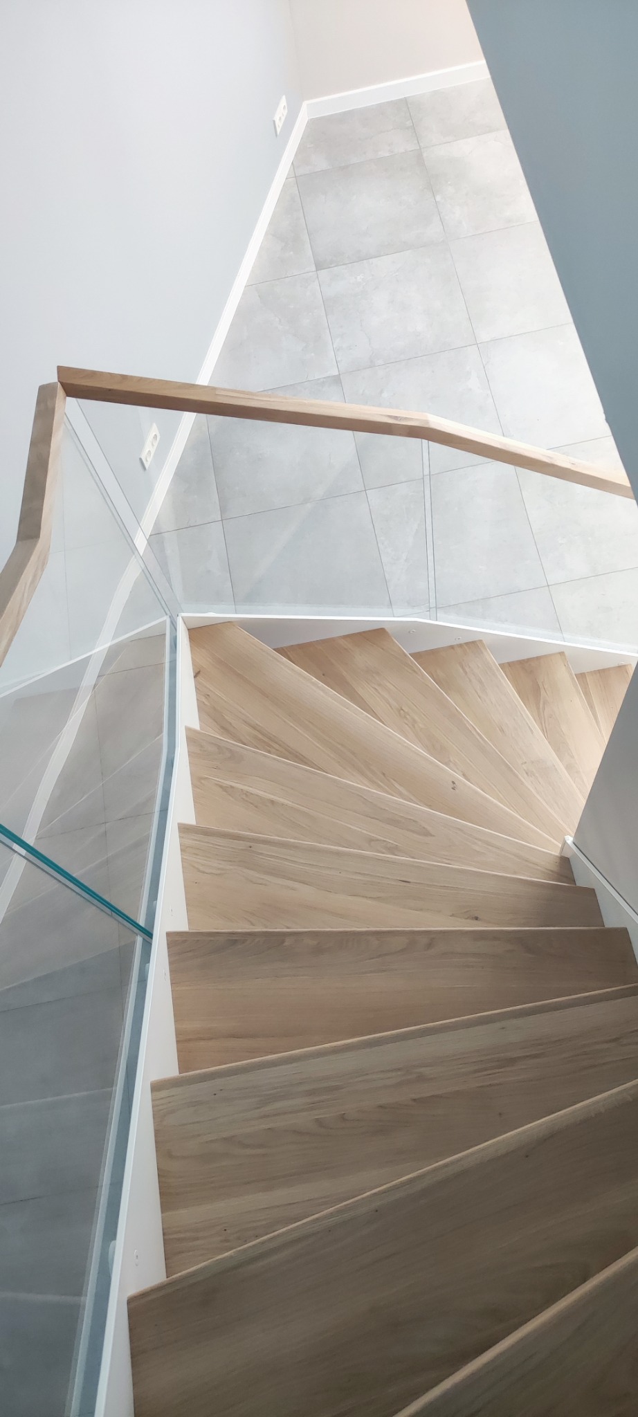 Kaskadowe schody z jasnego drewna z przezroczystą balustradą szklaną i drewnianą poręczą, widok z góry na szare płytki podłogowe.