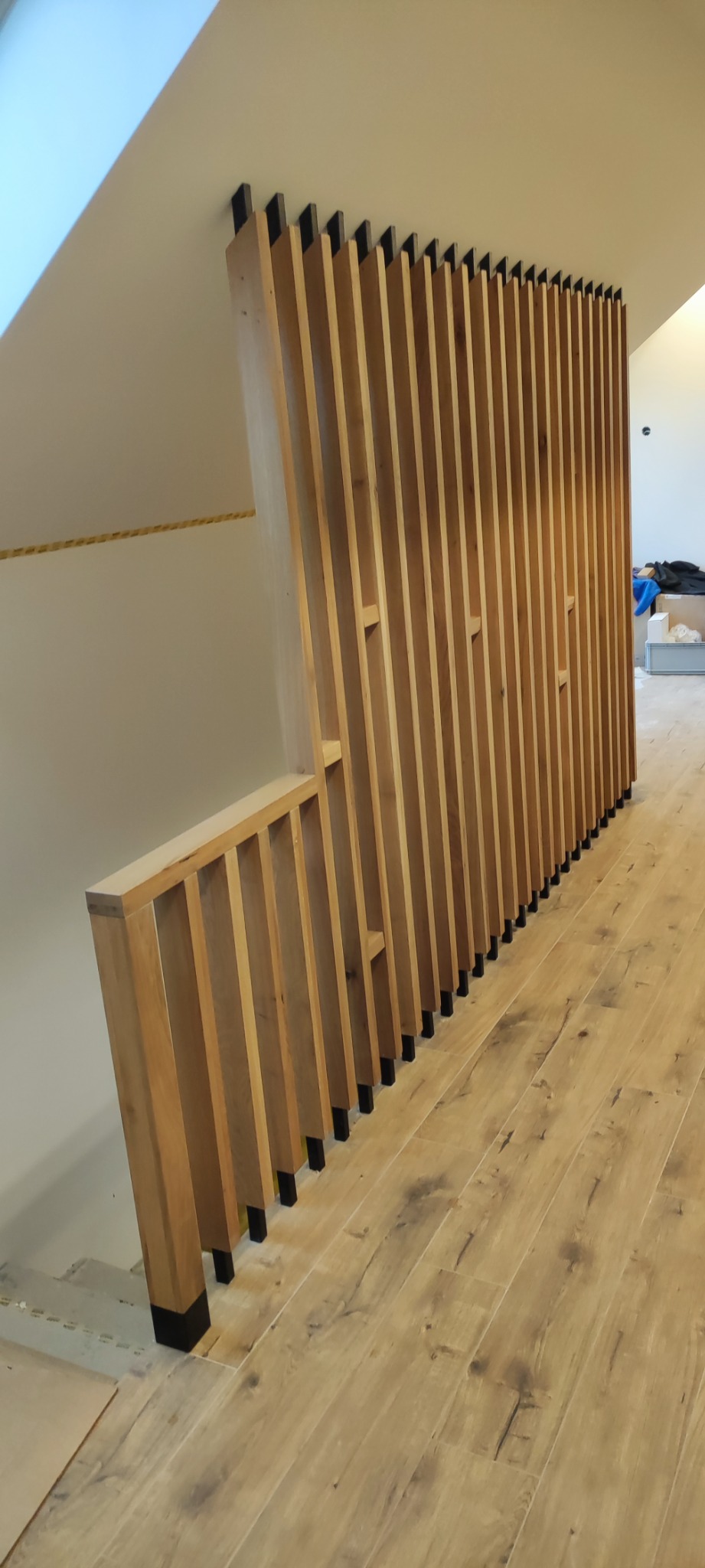 Drewniana balustrada schodowa z pionowymi szczeblami i czarnymi akcentami, oddzielająca przestrzeń na poddaszu. Jasna podłoga imitująca drewno.