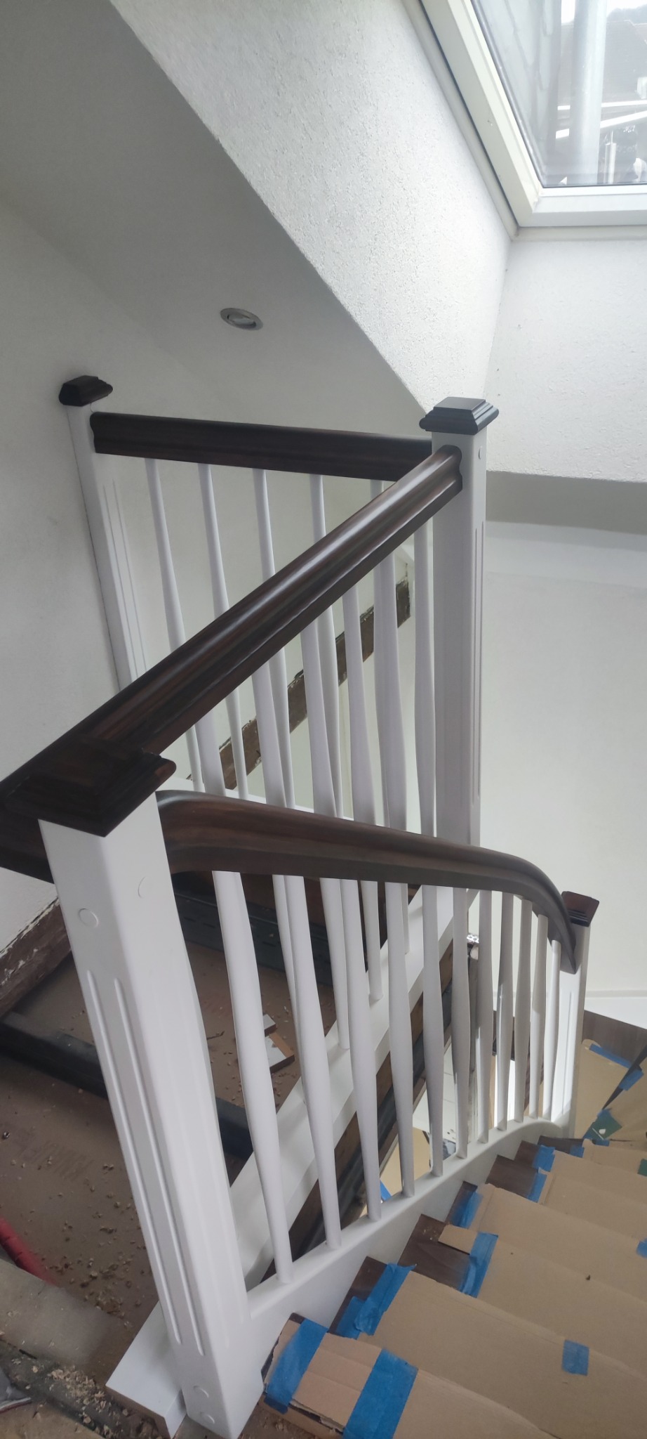 Wnętrze z widokiem na białe schody z ciemnobrązową poręczą, zabezpieczone kartonem na czas remontu. Ujęcie z góry, widoczne okno dachowe.