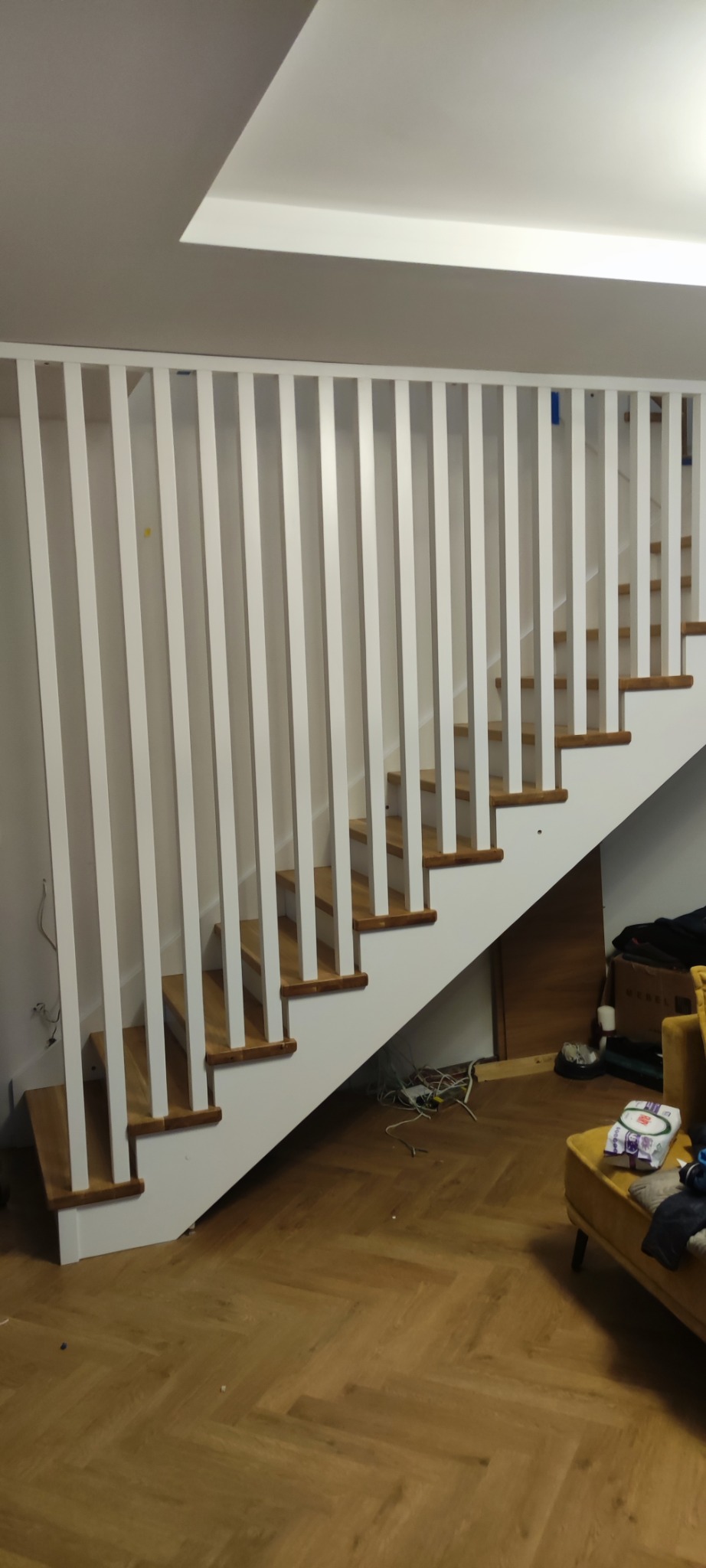 Nowoczesne schody drewniane z białą balustradą z pionowych listew. Parkiet w jodełkę na podłodze. Widoczne okablowanie i elementy wyposażenia wnętrza.