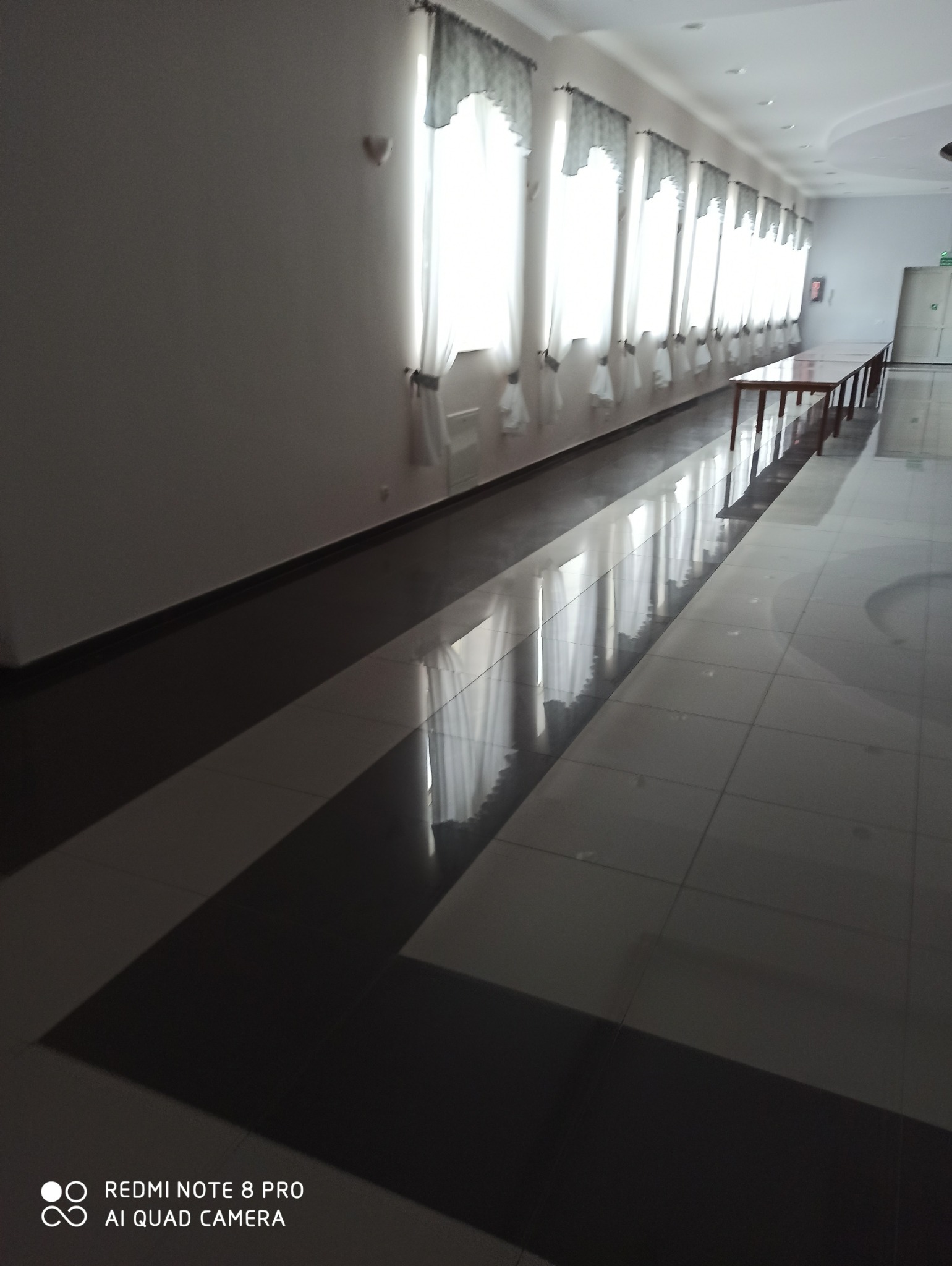 Długi korytarz z rzędem okien z dekoracyjnymi firanami. Na podłodze błyszczące, ciemne płytki z jasnymi pasami. W tle widoczne stoły i drzwi.