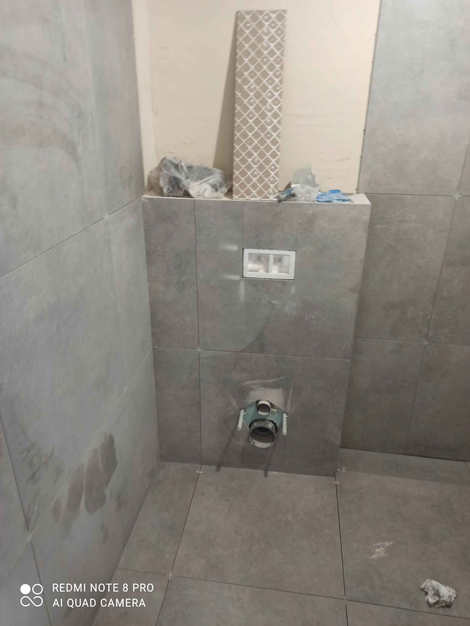 Szara, matowa glazura w łazience w trakcie montażu. Widoczny stelaż podtynkowy WC i fragment rury odpływowej. Na górze ściany leży płytka dekoracyjna.