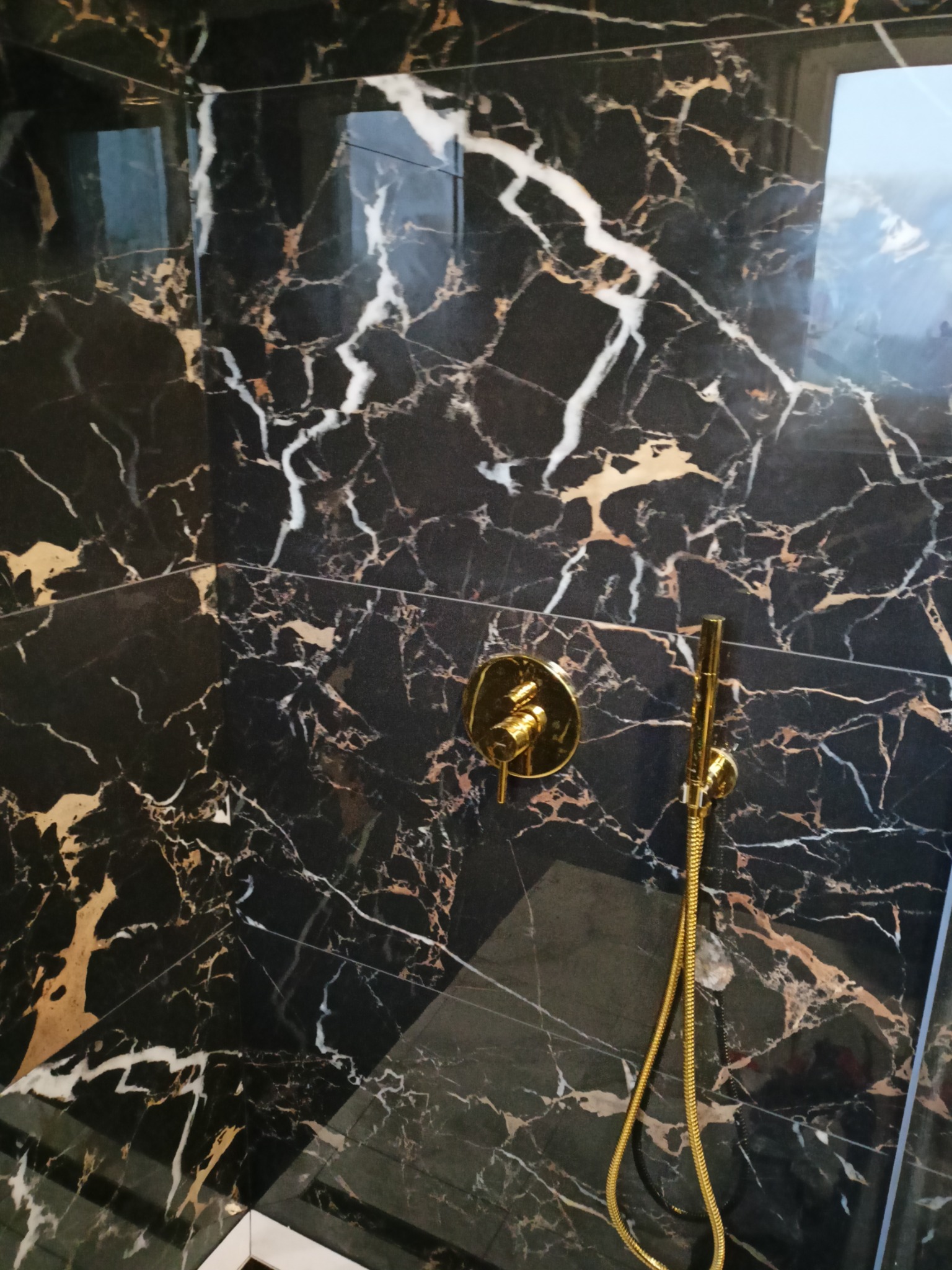 Wykończona kabina prysznicowa z czarnymi płytkami imitującymi marmur z białymi i złotymi żyłami oraz złotą armaturą. Nowoczesny design łazienki.