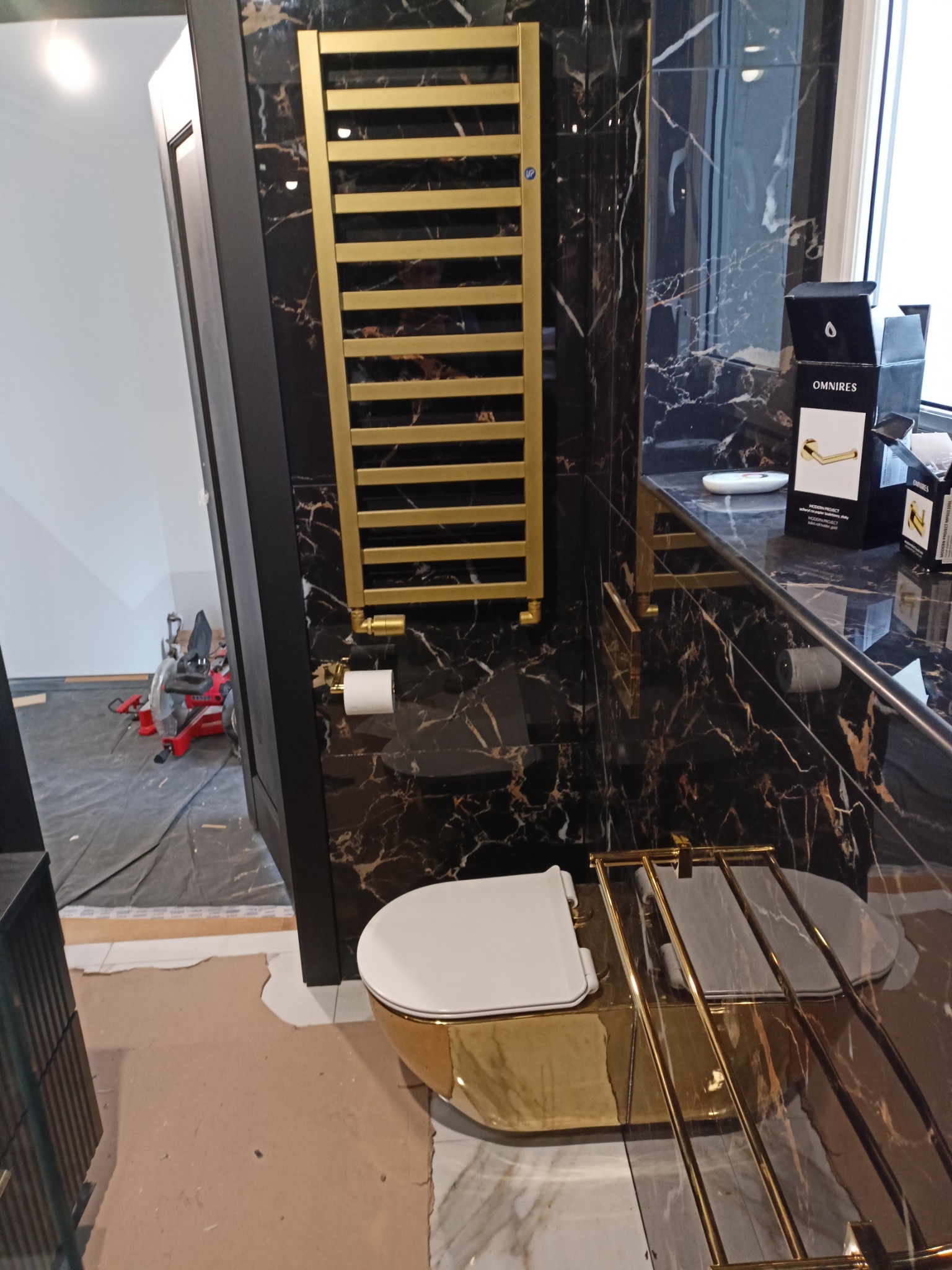 Luksusowa łazienka w trakcie wykończenia: złota armatura, marmurowe ściany z wyraźnym użyleniem. Na podłodze widoczne ślady prac wykończeniowych, narzędzia w tle.