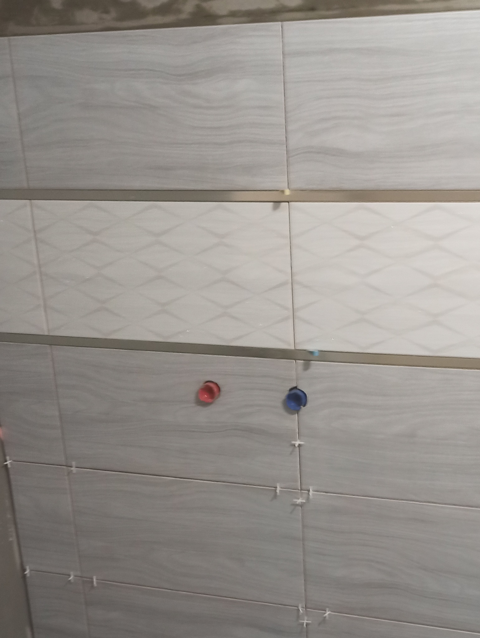 Fragment ściany z ułożonymi płytkami w odcieniach szarości i bieli, widoczne krzyżyki dystansowe i otwory na instalację wodną.