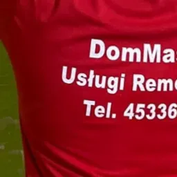 Czerwona koszulka z białym napisem 'DomMaster Usługi Remontowe Tel. 453369851', na tle zielonej, częściowo odrapanej ściany z cegieł.
