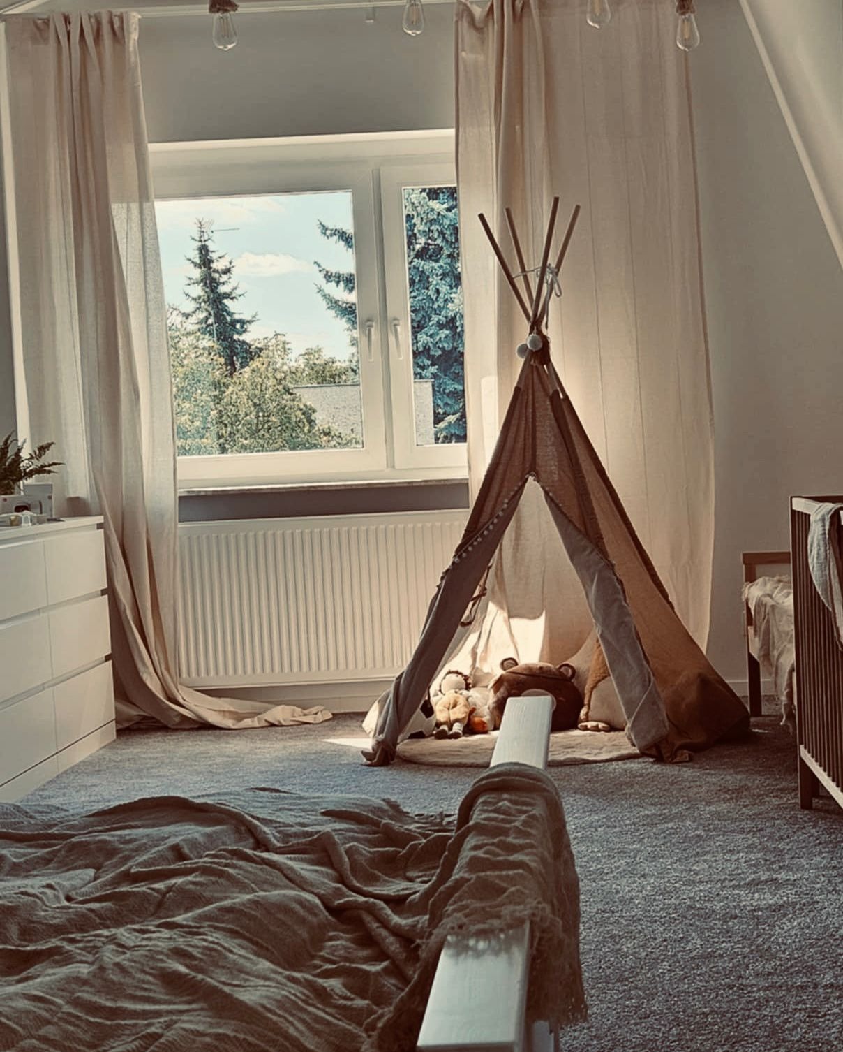 Jasny pokój dziecięcy z tipi, zasłonami i widokiem na zieleń za oknem. Ustawne wnętrze z łóżeczkiem i komodą. Stonowana kolorystyka, naturalne światło.
