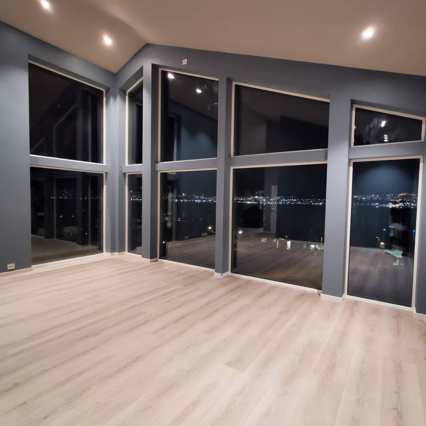 Nowoczesne wnętrze z dużymi oknami i widokiem na nocne miasto. Jasna podłoga, szare ściany, minimalistyczny design. Oświetlenie sufitowe podkreśla przestrzeń.