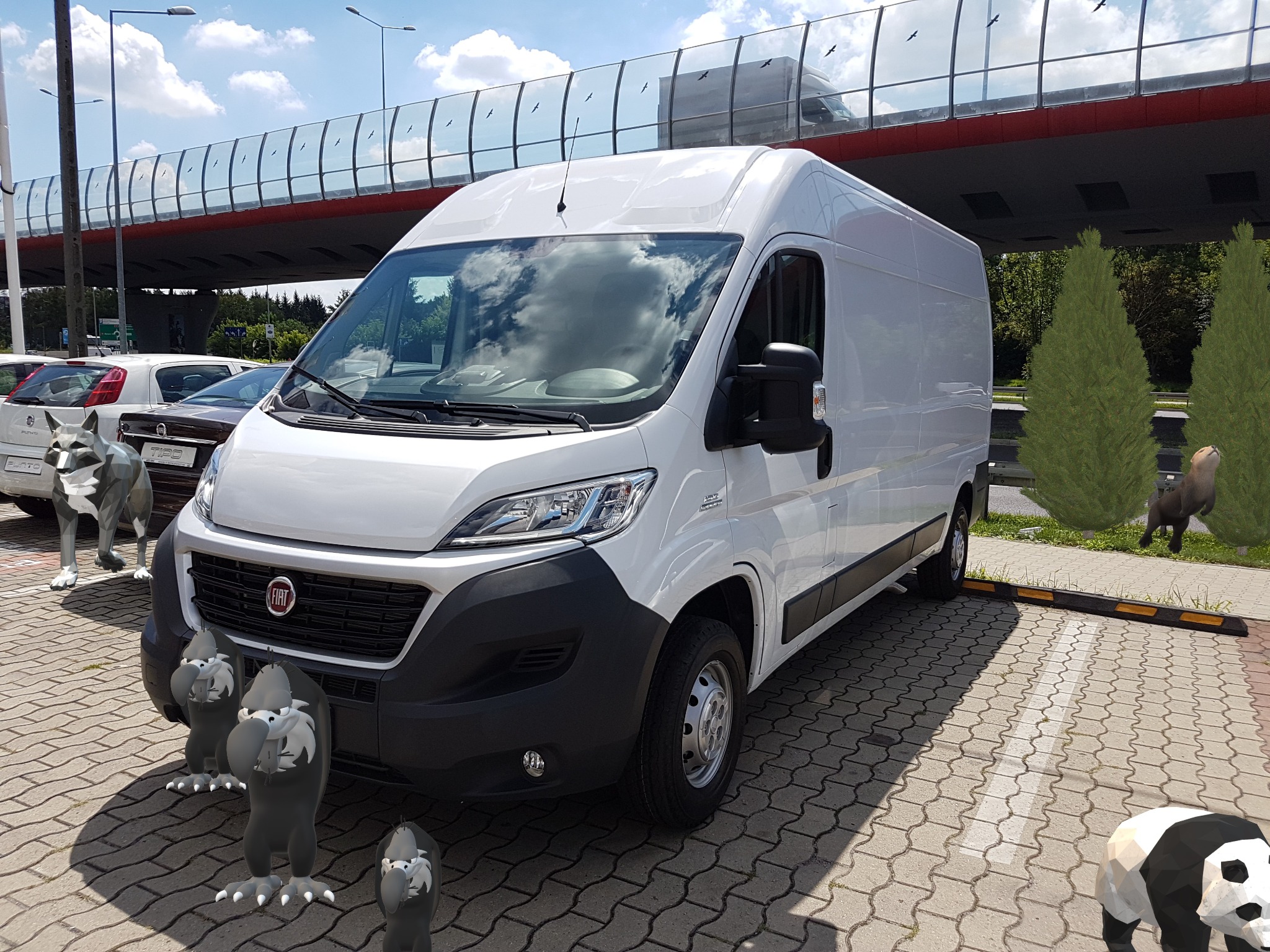 Biały furgon Fiat Ducato zaparkowany na brukowanym parkingu z dodatkami graficznymi: figurki zwierząt, drzewa i odbicie innego pojazdu na wiadukcie w szybie.