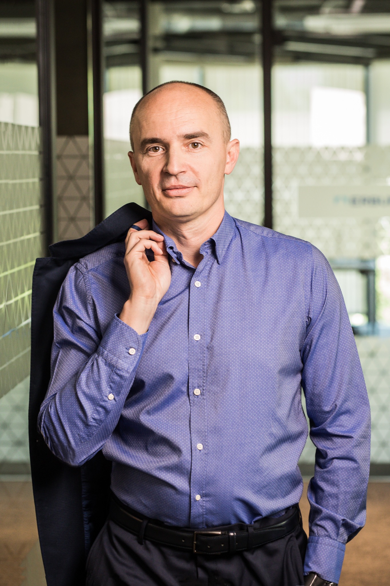 Mężczyzna w niebieskiej koszuli z marynarką na ramieniu, stoi na tle biura. Uśmiechnięty, patrzy wprost. Profesjonalny wizerunek.