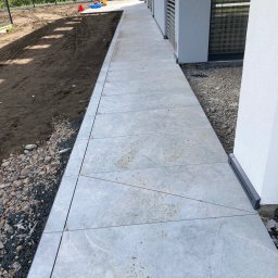 RYŚ - Ułożona ścieżka z jasnych, kwadratowych płyt chodnikowych prowadząca wzdłuż budynku z szarymi roletami, obok ziemi i kamieni.