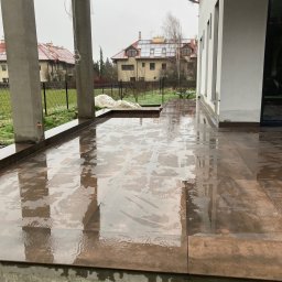 RYŚ - Świeżo ułożone, mokre płytki na zewnątrz domu, z widocznymi słupami i fragmentem budynku w tle. Płytki w odcieniach brązu, odbijające światło.