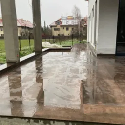 Świeżo ułożone, mokre płytki na zewnątrz domu, z widocznymi słupami i fragmentem budynku w tle. Płytki w odcieniach brązu, odbijające światło.