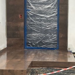 RYŚ - Błyszczące, brązowe płytki na schodach wejściowych do budynku, z zabezpieczonym folią wejściem. Widoczna instalacja elektryczna i fragment taśmy ostrzegawczej.