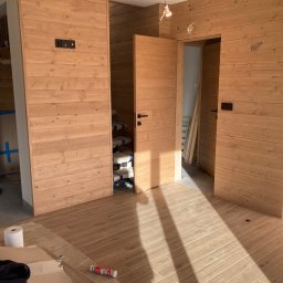 RYŚ - Wnętrze z wykończonymi drewnianymi ścianami i podłogą z paneli imitujących drewno. Widoczne otwarte drzwi i narzędzia, co sugeruje etap prac wykończeniowych w nowym lub remontowanym pomieszczeniu.