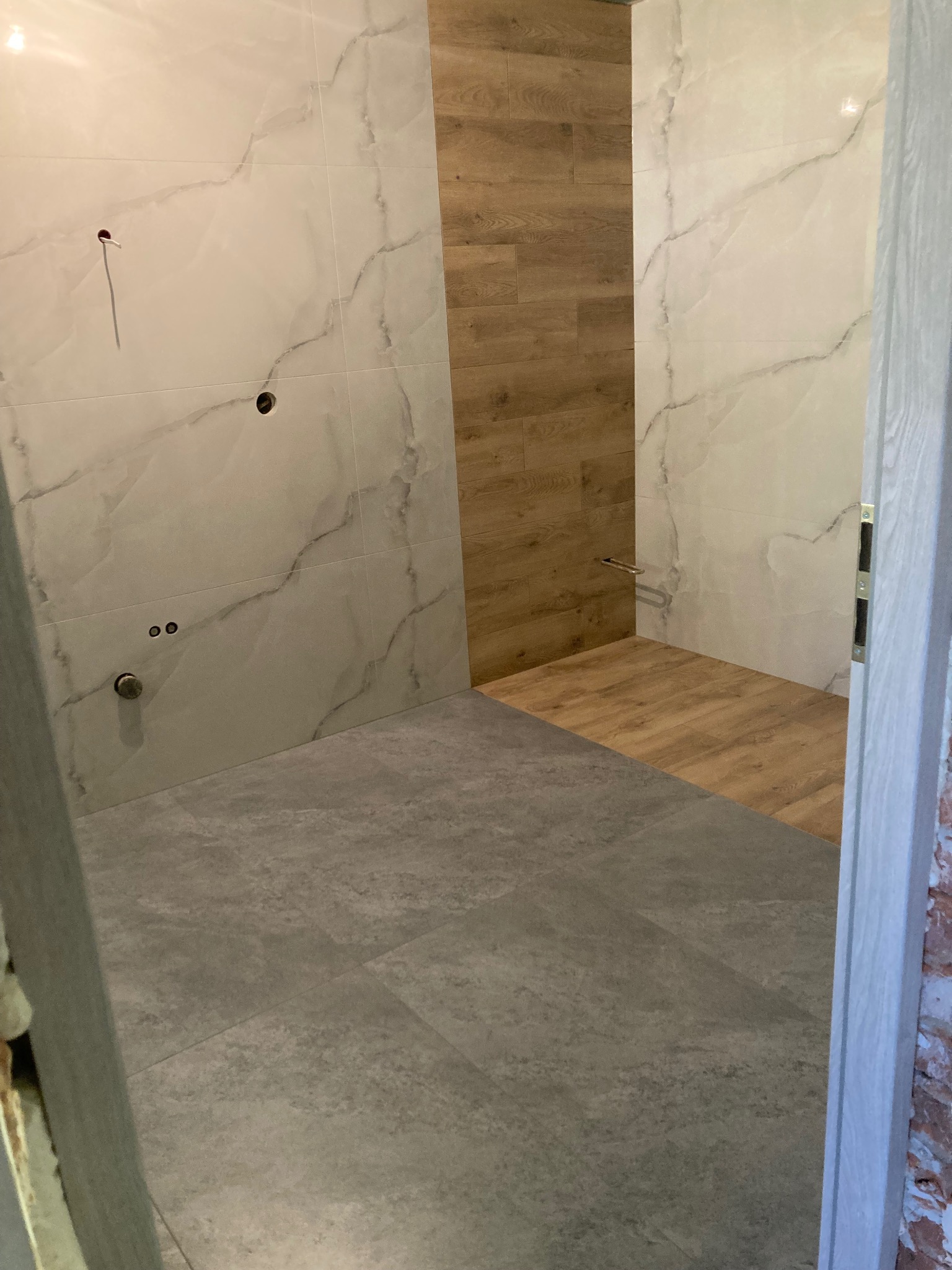 Wykończona łazienka z szarymi płytkami podłogowymi i marmurowymi na ścianach, oraz drewnopodobnymi w strefie prysznica. Widoczne wyprowadzenia instalacji.