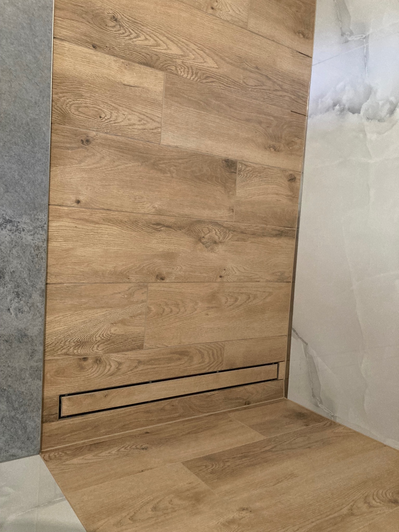 Wykończona kabina prysznicowa z odpływem liniowym i okładziną imitującą drewno na ścianie i podłodze, sąsiadująca z szarą i białą płytką.