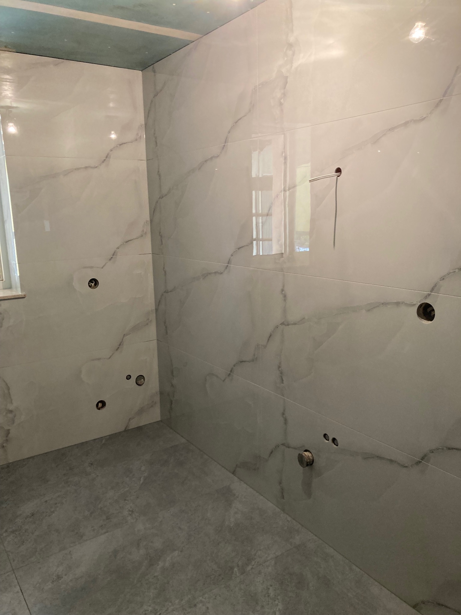 Wykończona łazienka z połyskującymi, marmurkowymi płytkami na ścianach i szarymi płytkami podłogowymi. Widoczne otwory na armaturę i odbicie okna w płytkach.