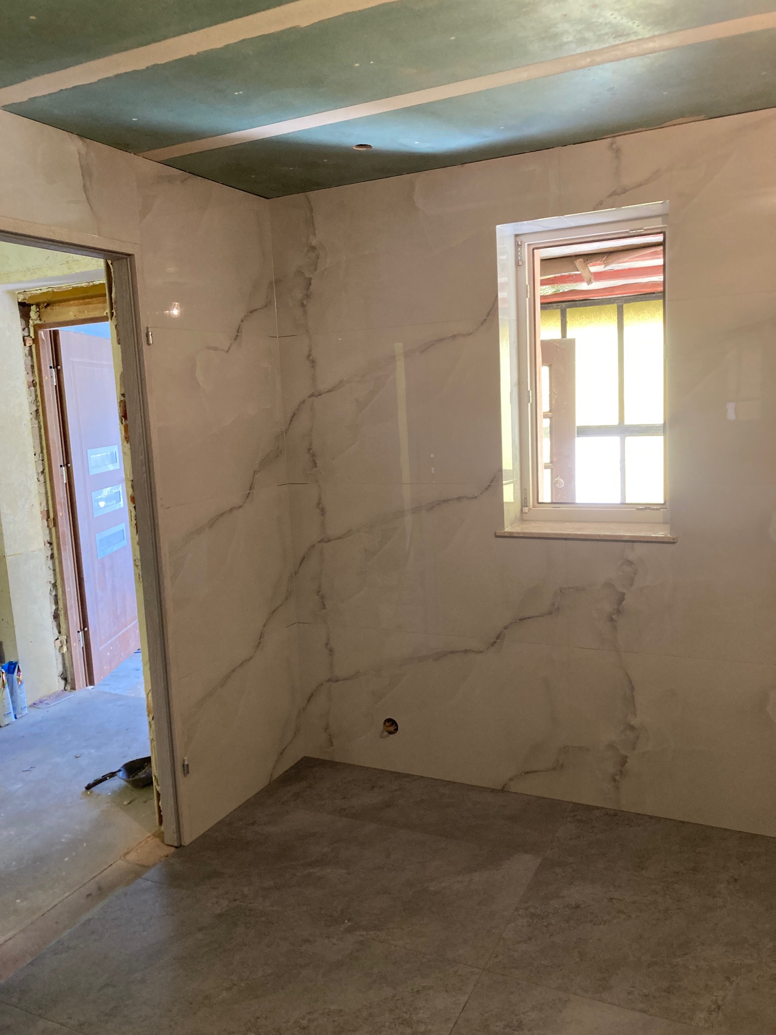 Wykończona łazienka z płytkami imitującymi marmur na ścianach i szarą podłogą. Widoczne okno i surowy sufit. Drzwi bez ościeżnicy.