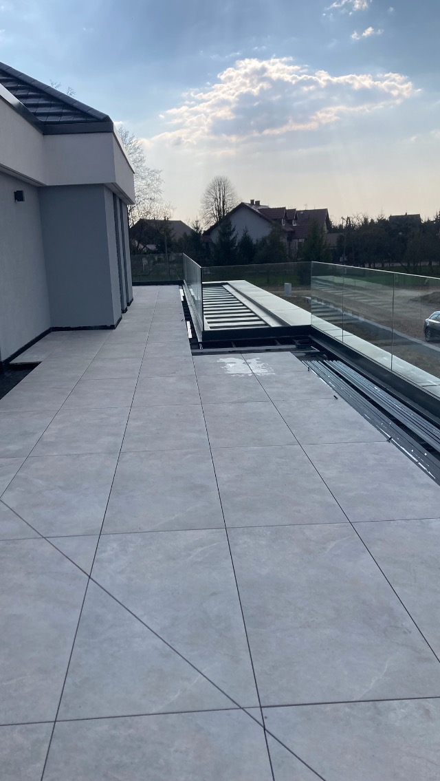 Jasnoszara, kwadratowa płytka na balkonie z nowoczesną szklaną balustradą. W tle widoczne budynki mieszkalne i błękitne niebo z chmurami.