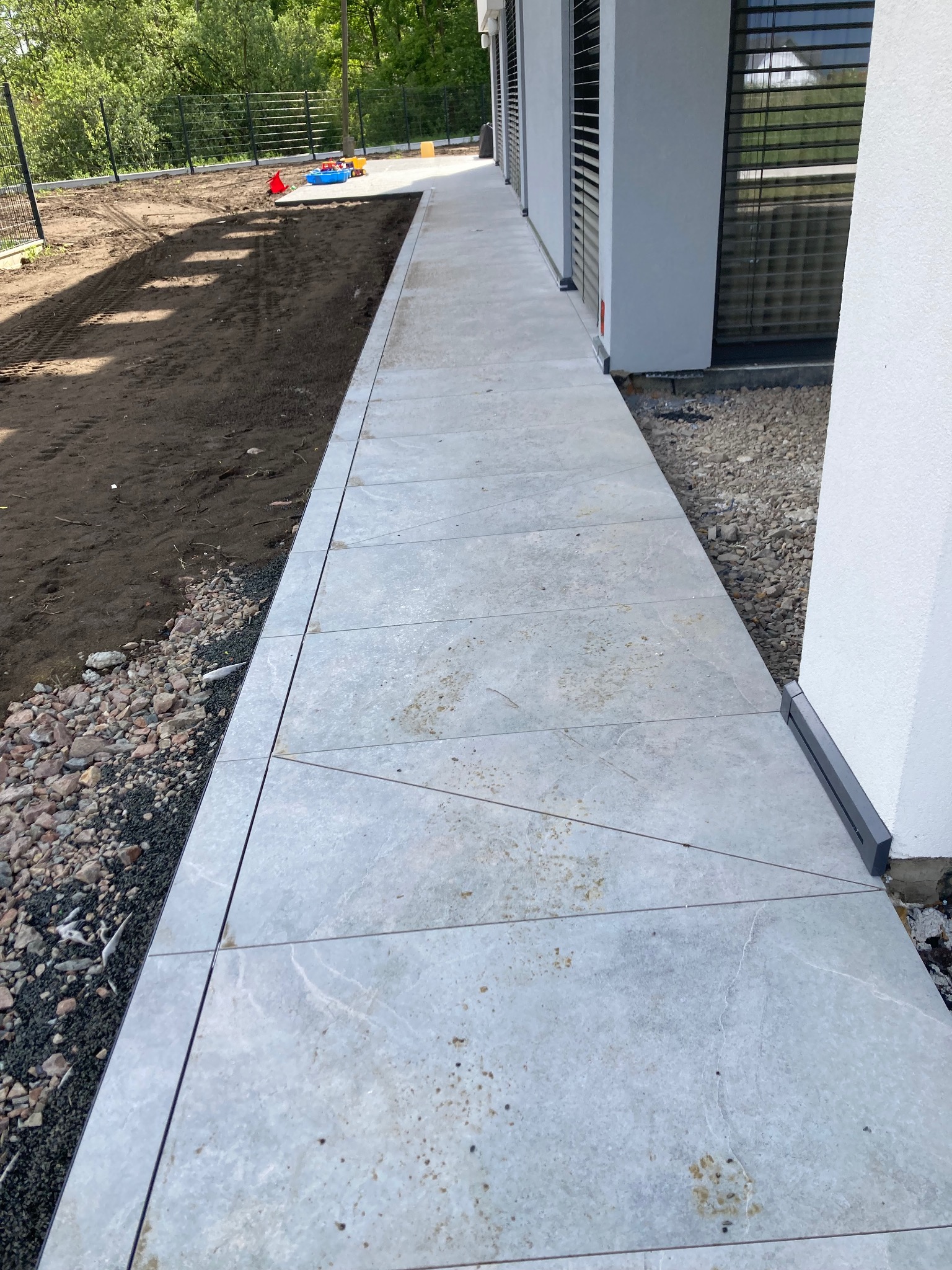 Ułożona ścieżka z jasnych, kwadratowych płyt chodnikowych prowadząca wzdłuż budynku z szarymi roletami, obok ziemi i kamieni.