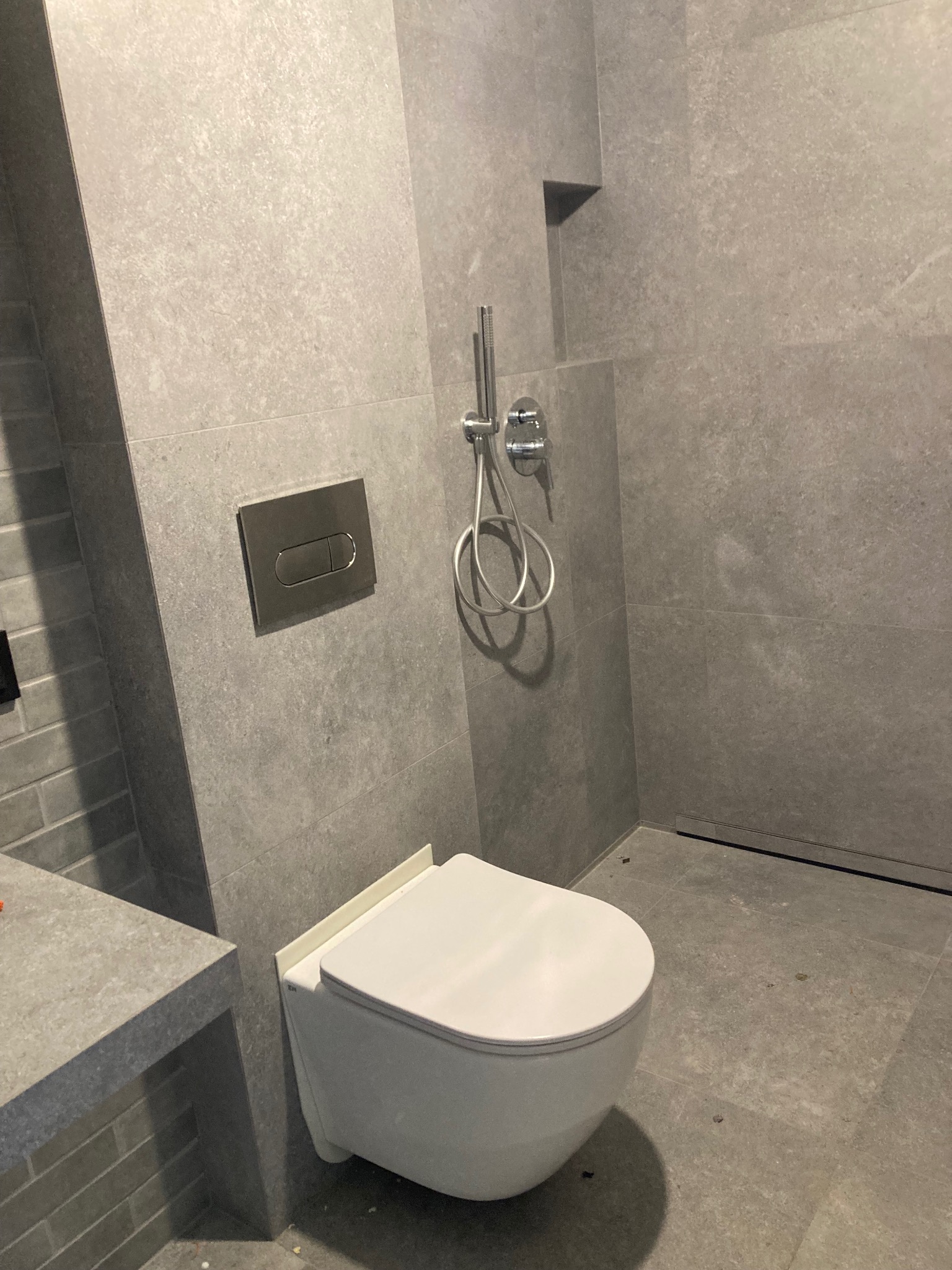 Nowoczesna łazienka z podtynkową toaletą i deszczownicą. Szare płytki imitujące kamień, minimalistyczny design. Widoczny odpływ liniowy.