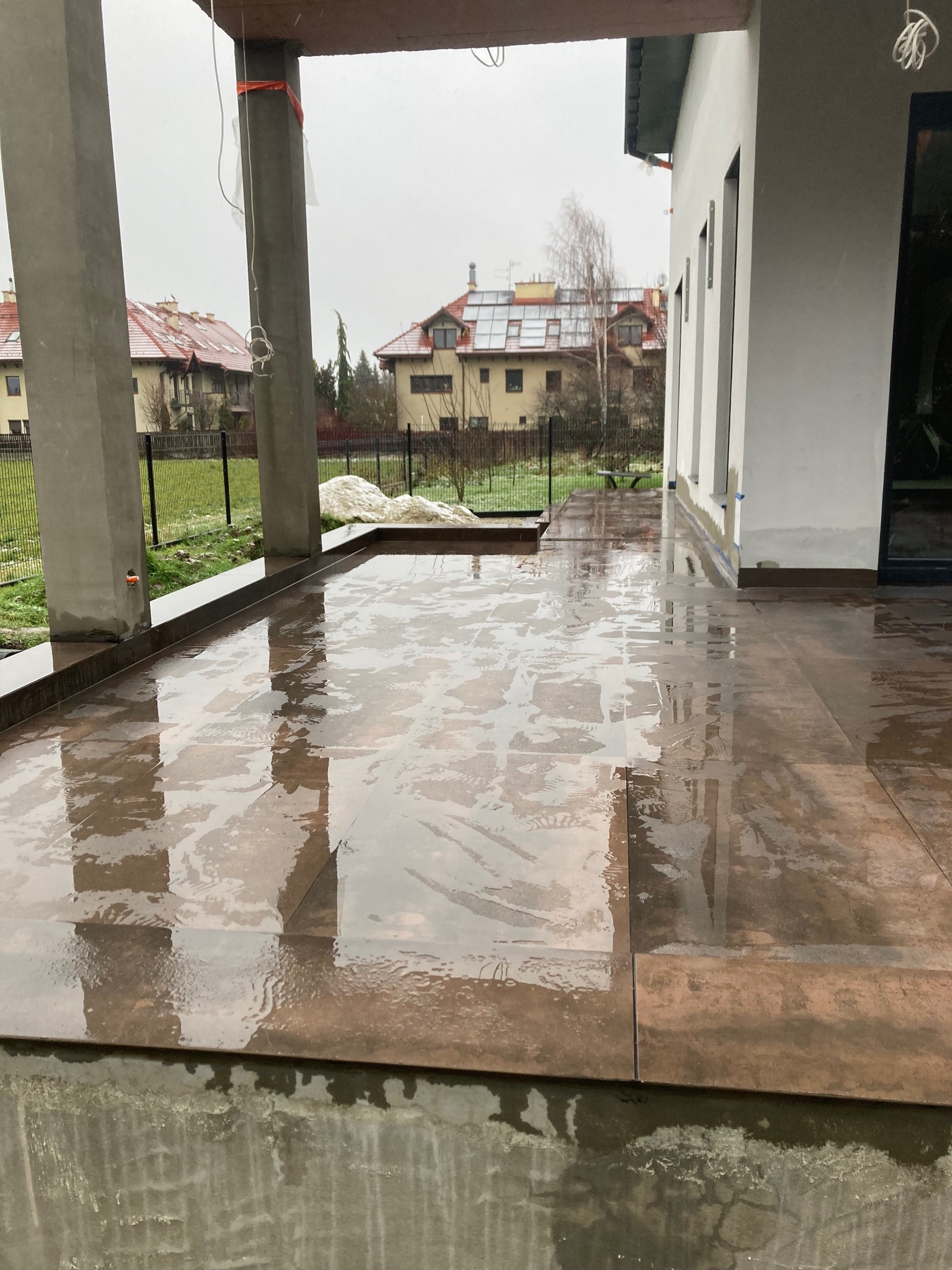 Świeżo ułożone, mokre płytki na zewnątrz domu, z widocznymi słupami i fragmentem budynku w tle. Płytki w odcieniach brązu, odbijające światło.