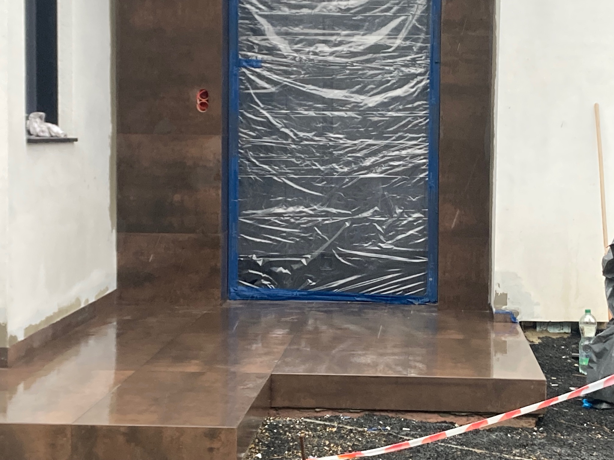 Błyszczące, brązowe płytki na schodach wejściowych do budynku, z zabezpieczonym folią wejściem. Widoczna instalacja elektryczna i fragment taśmy ostrzegawczej.